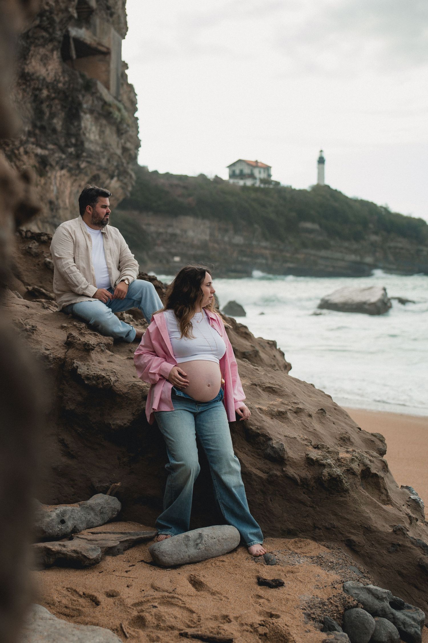 Pregnant couple posing on rocky coastal cliffs with a lighthouse and ocean waves in the background.