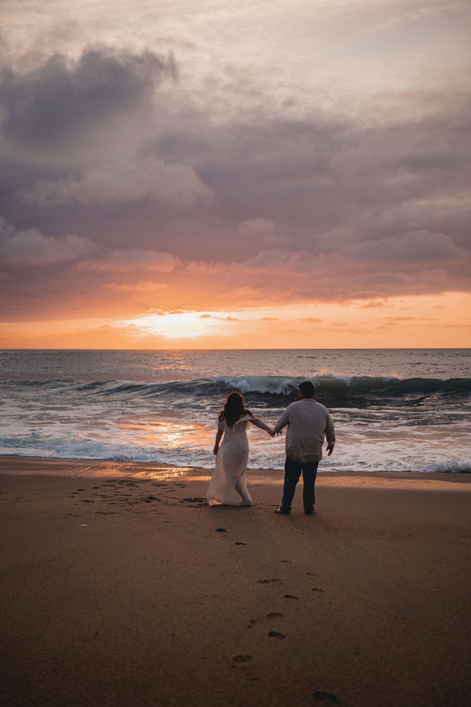 Couple walking hand-in-hand along a sandy beach at a stunning golden sunset, with footprints trailing behind.