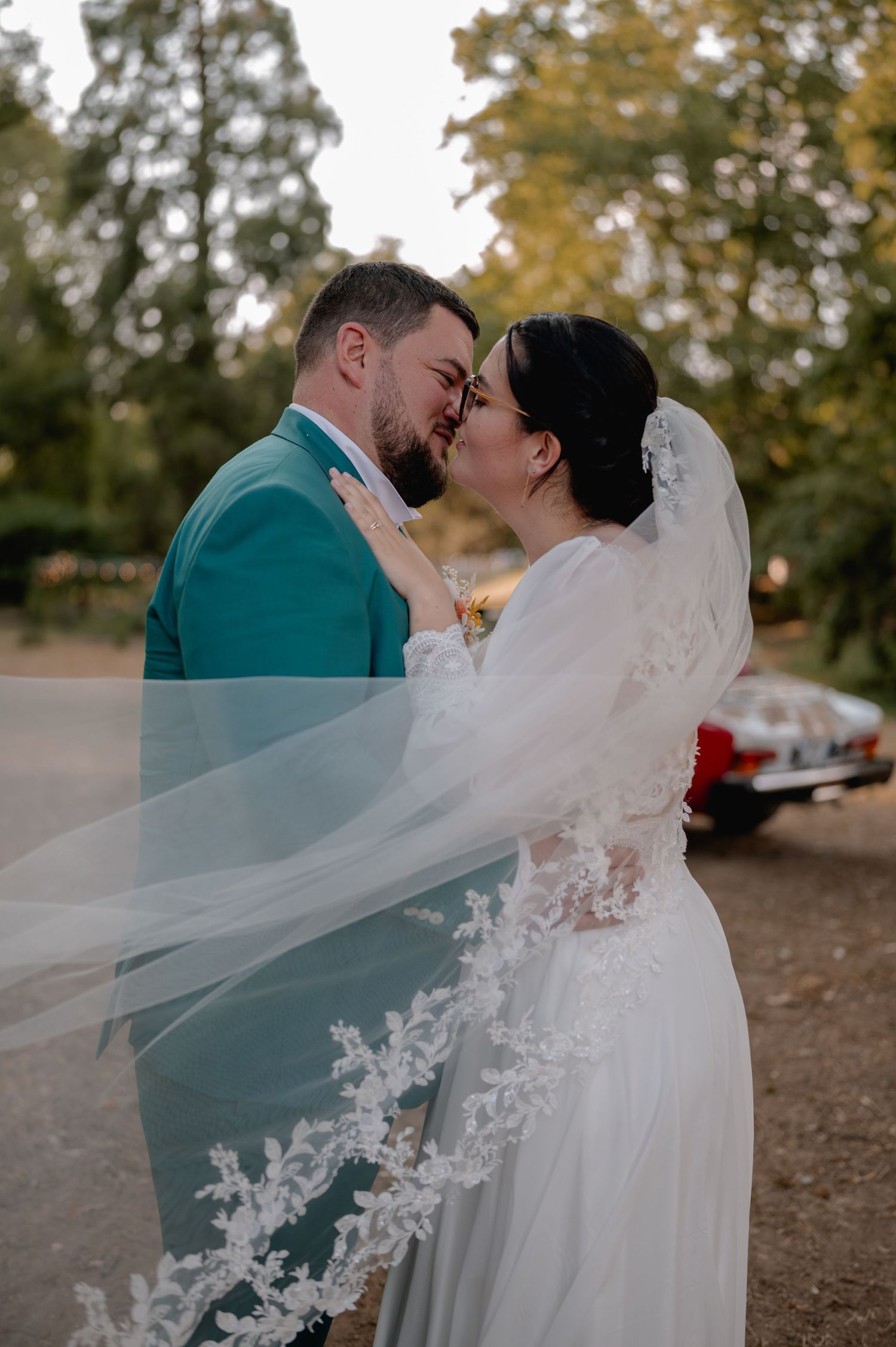 Bride in lace veil and groom in teal suit share intimate moment outdoors.