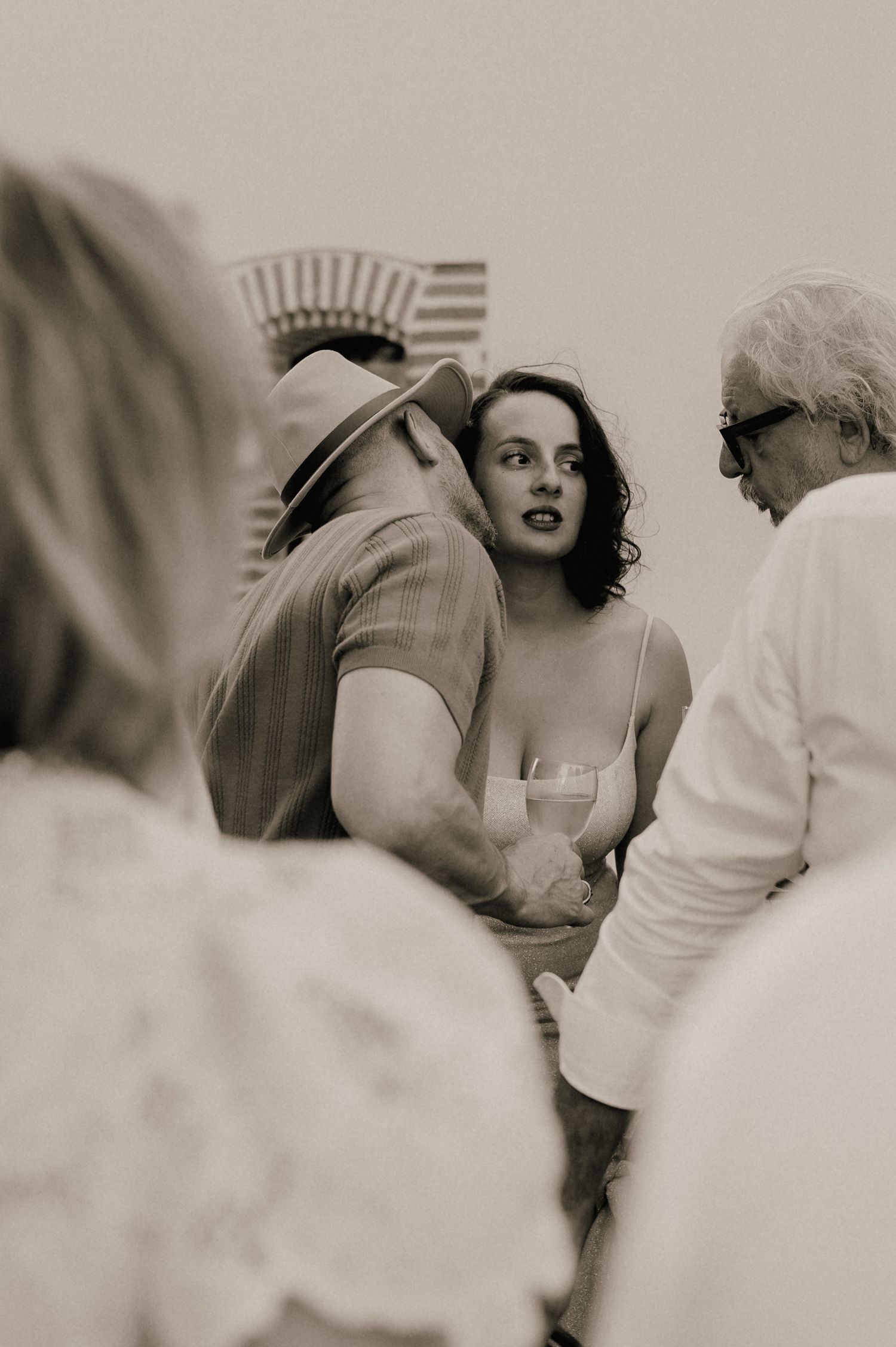 Black and white photo of a woman in a spaghetti strap dress holding a wine glass at an outdoor social gathering.