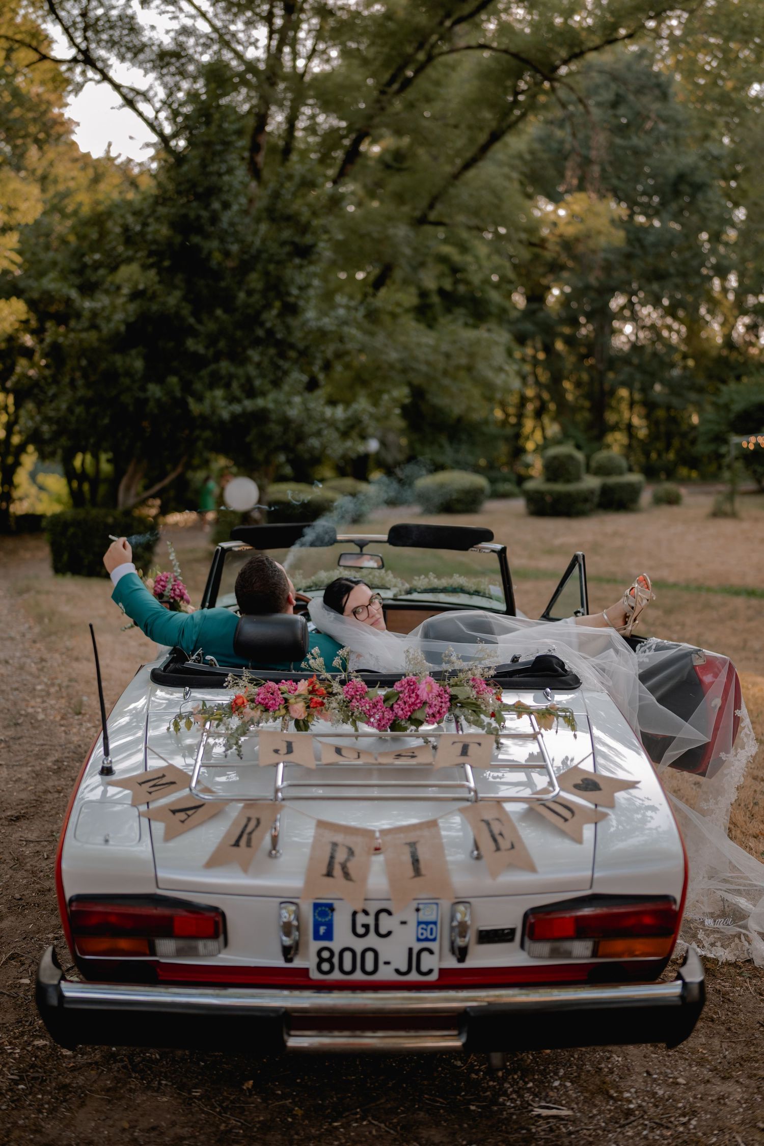 Newlyweds sitting in a decorated white convertible car with floral arrangements and a MARIE banner at a wedding.