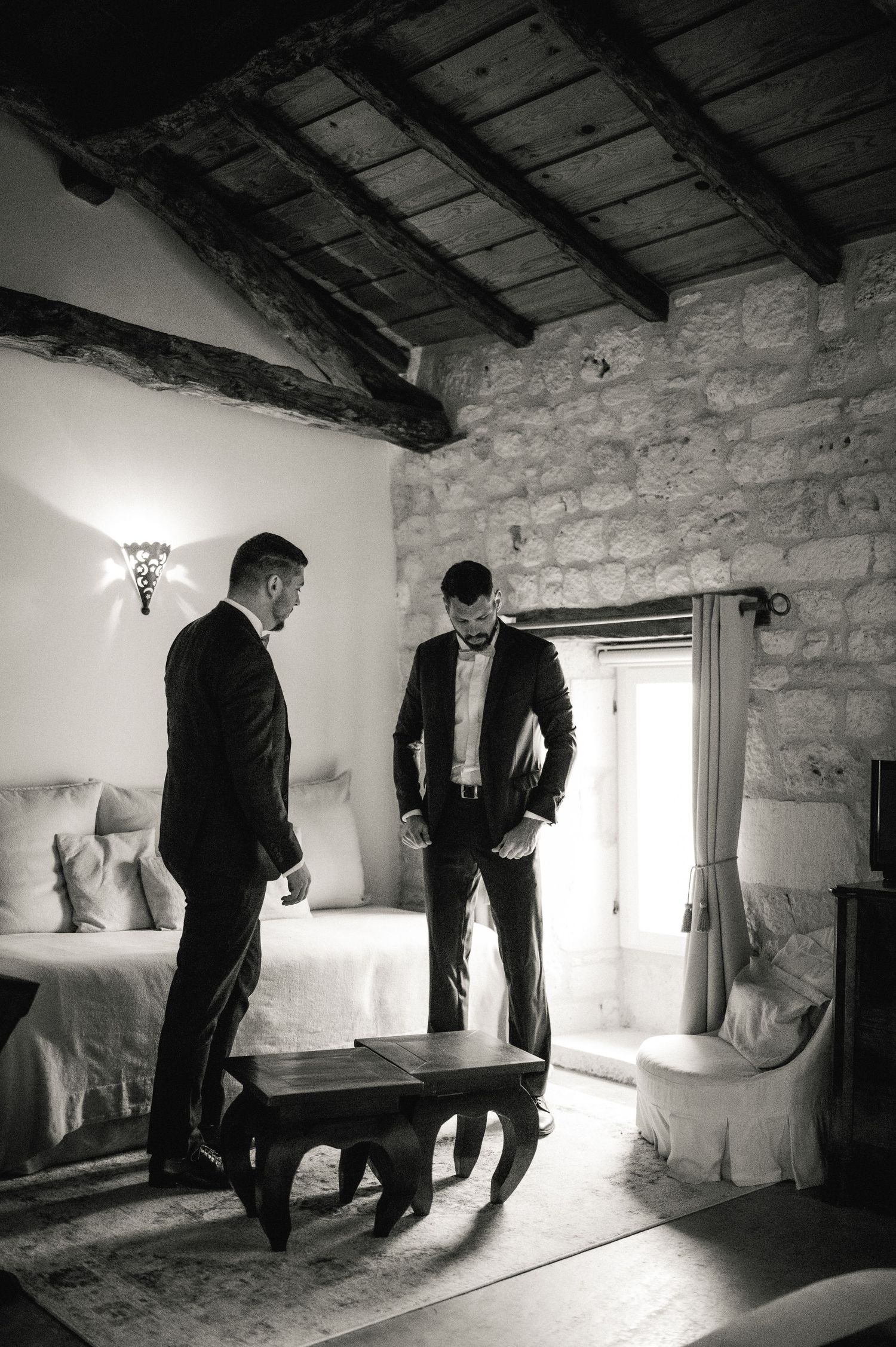 Black and white photo of two men in suits preparing for a wedding in rustic stone room.