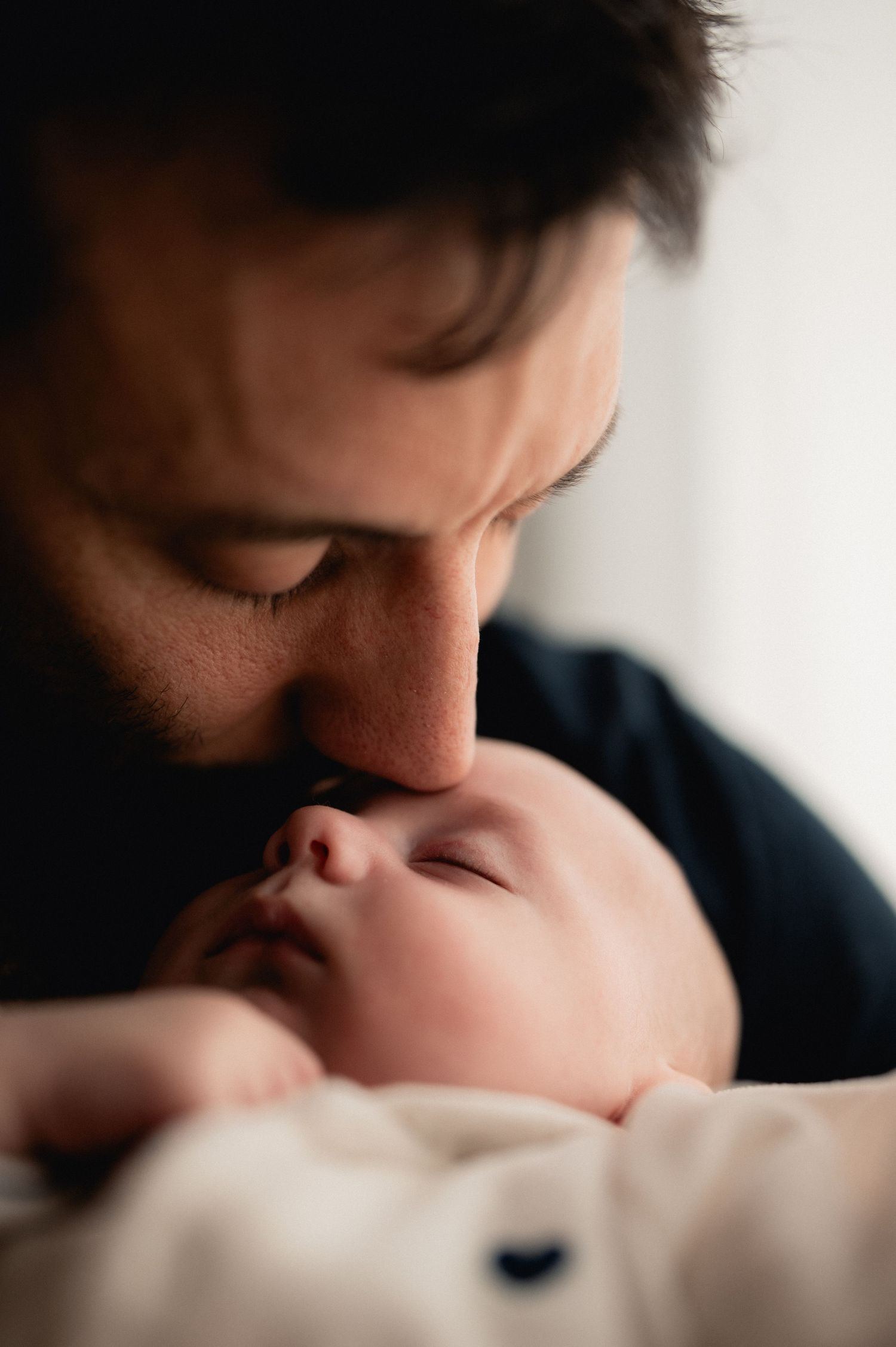 Father gently kissing his sleeping newborn baby on the nose in a tender close-up portrait.