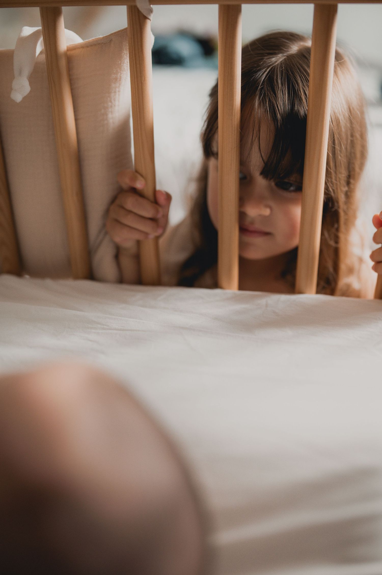 A curious young girl peers through wooden crib slats at a newborn baby lying inside the cot.