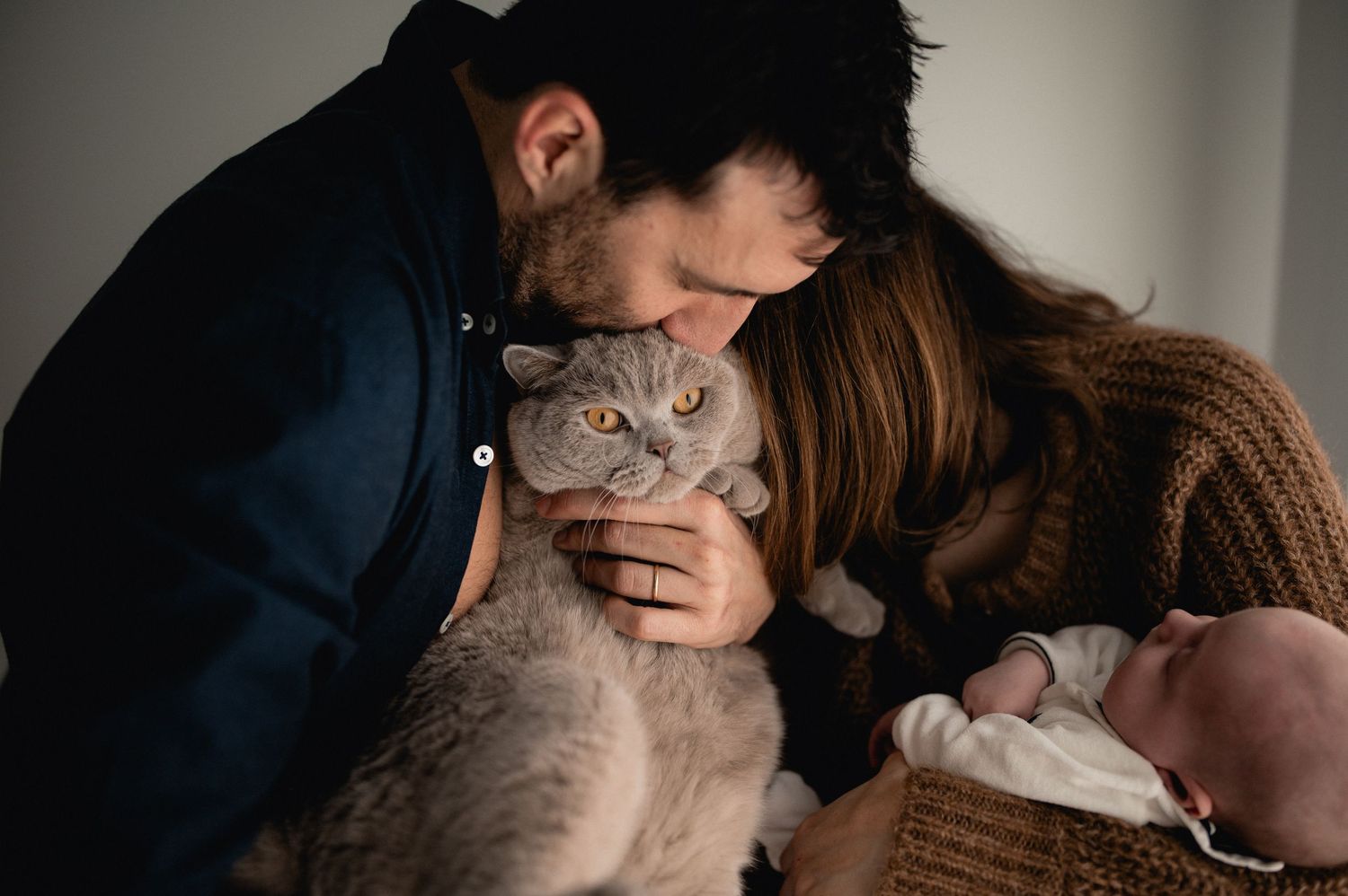 Couple kissing their fluffy grey cat while mother holds newborn baby in a wicker basket.