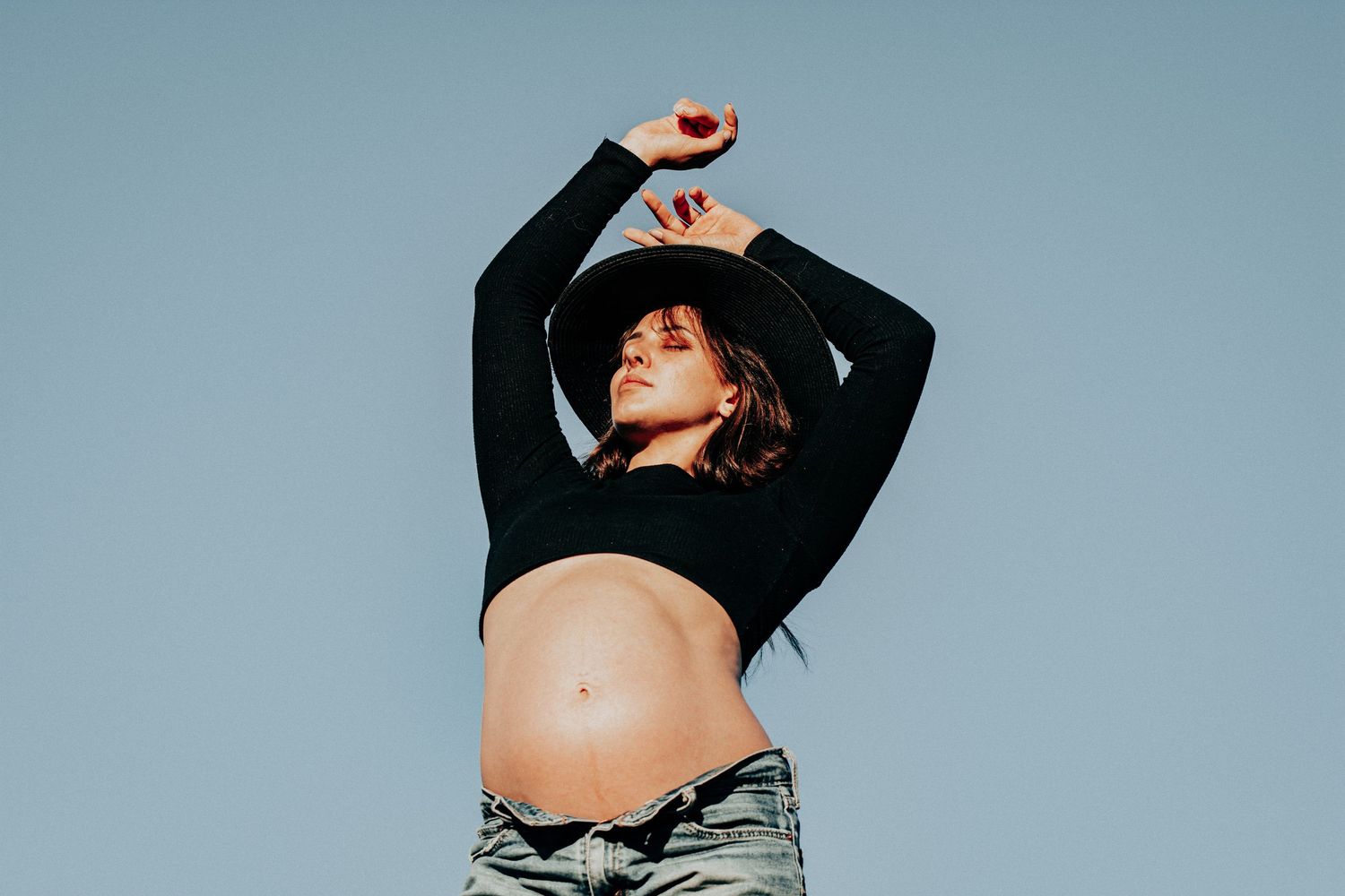 Pregnant woman in black crop top and hat poses against blue sky backdrop.