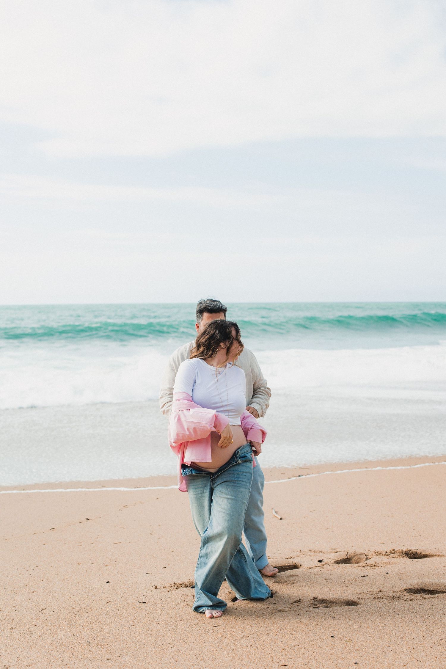 A couple embraces while walking barefoot on a sandy beach with turquoise ocean waves in the background.