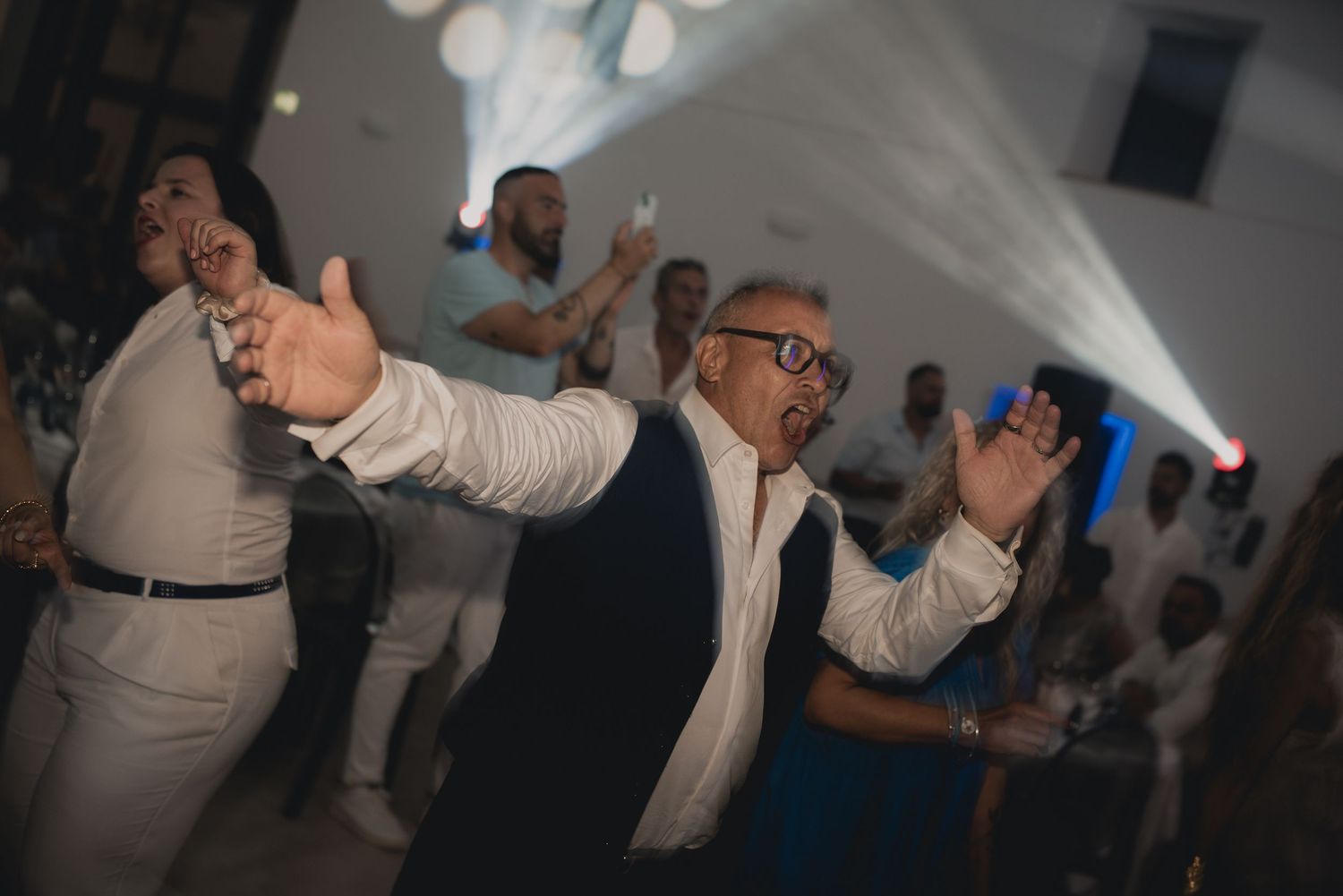 Man dancing energetically at a lively party event with bright stage lights and guests in white attire in the background.