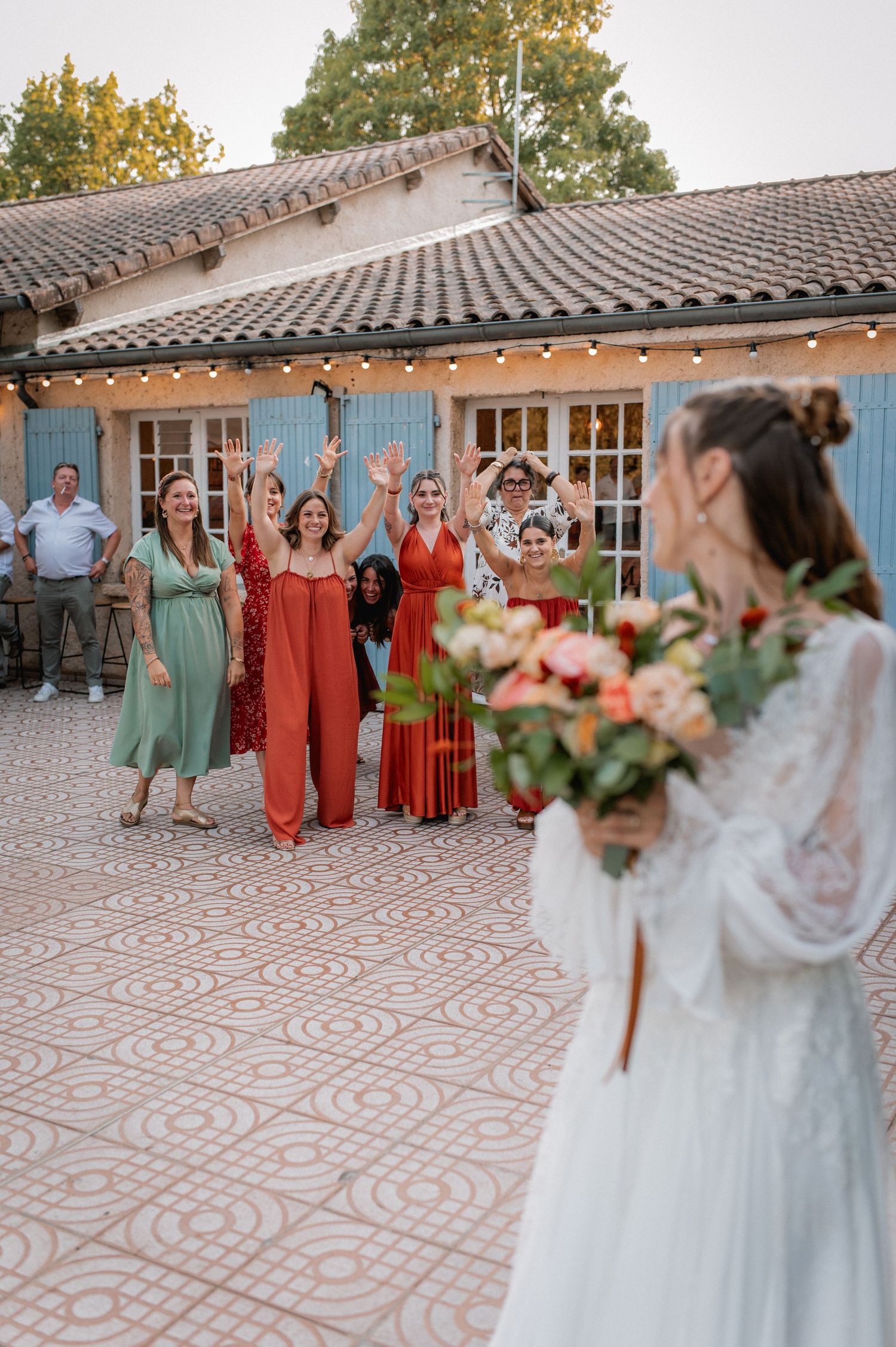 Bride preparing to toss bouquet to excited female wedding guests outside rustic venue with string lights.