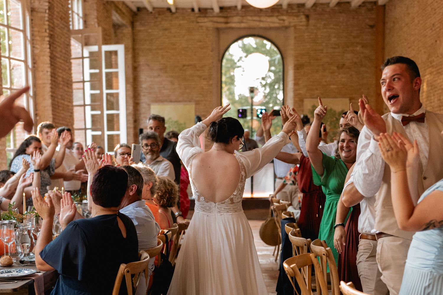Bride and groom enter wedding reception hall greeted by cheering guests in a rustic brick venue.