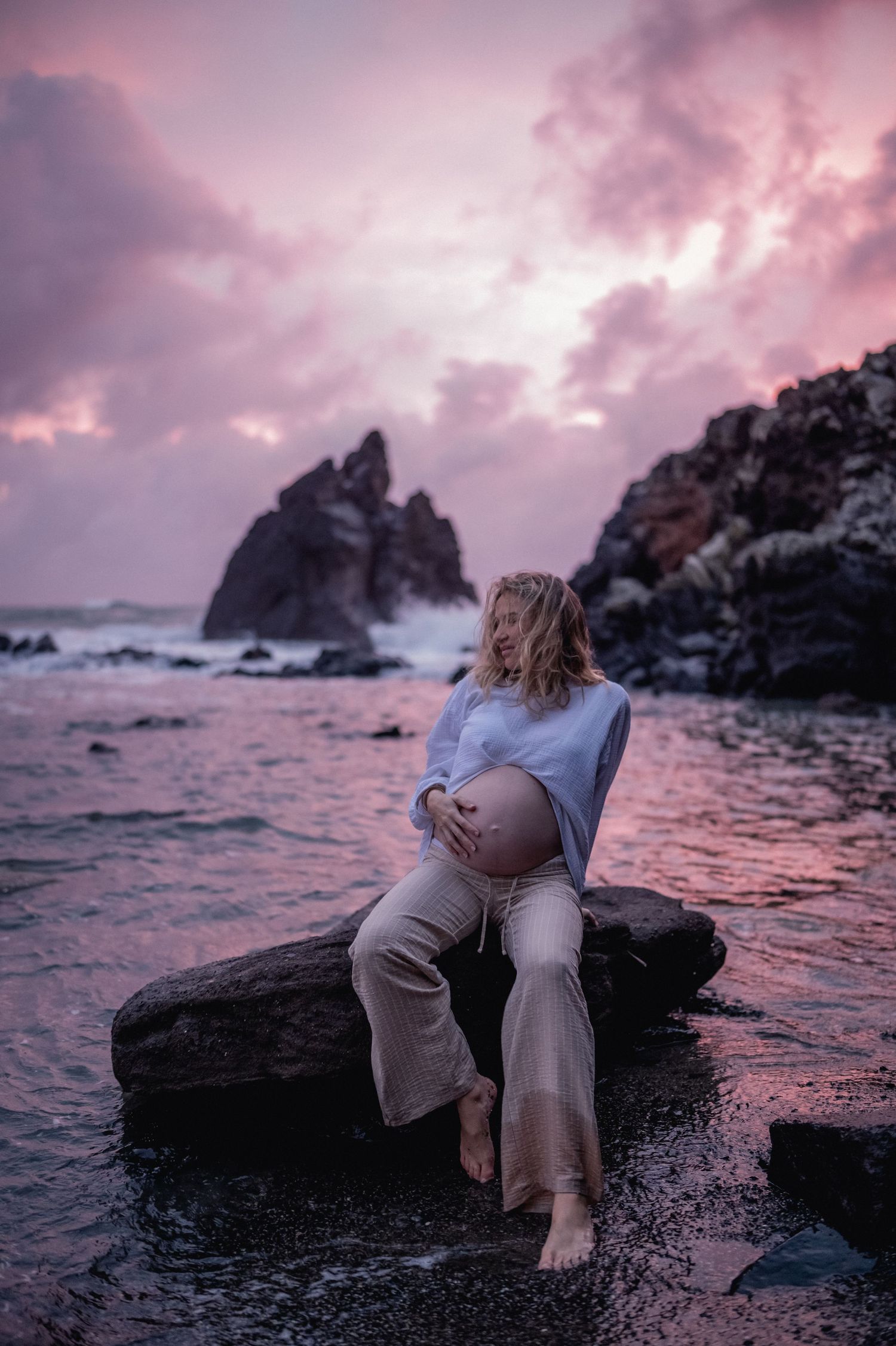 Pregnant woman sitting on rocks by ocean at pink sunset, cradling her baby bump barefoot.