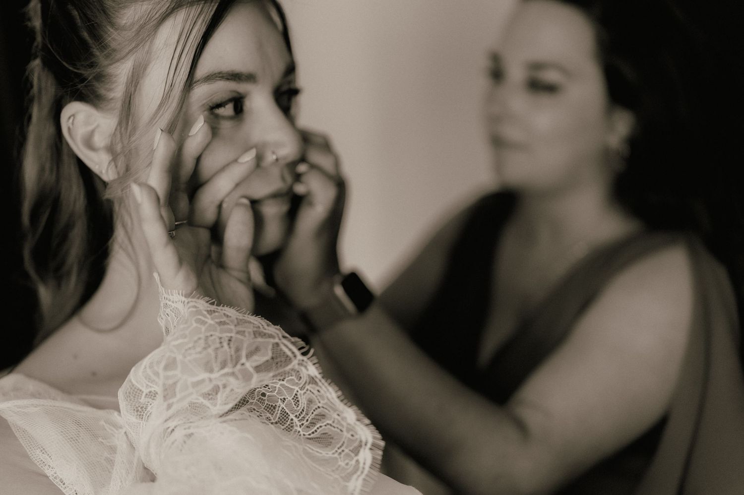 Black and white photo of emotional bride covering her mouth while bridesmaid fastens wedding dress buttons behind her.