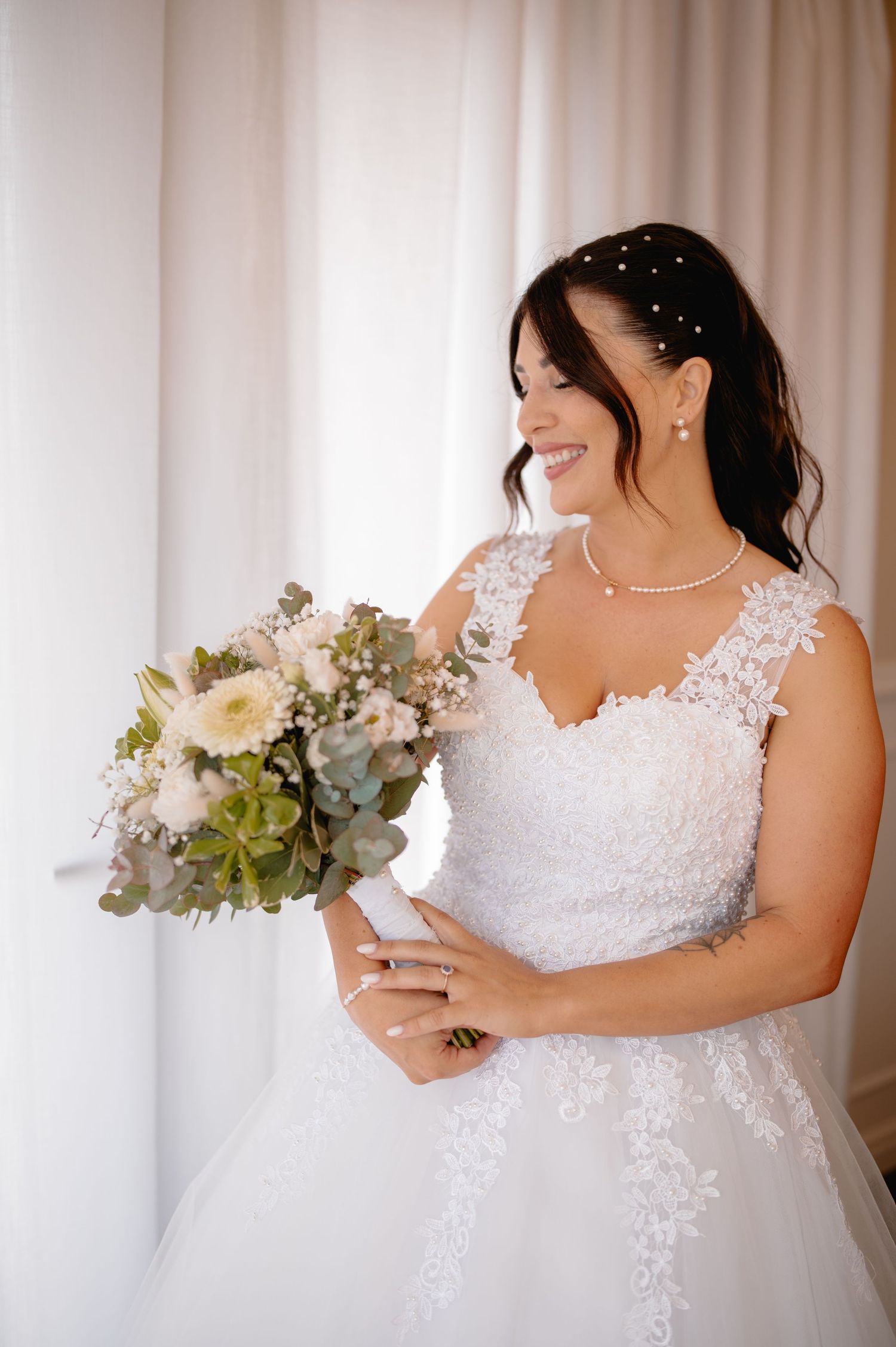 Smiling bride in white lace gown holding a bouquet of white and green flowers, wearing pearl jewelry.