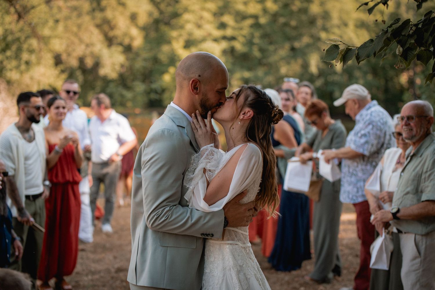 Bride and groom share first kiss at outdoor wedding ceremony surrounded by guests in warm golden light.