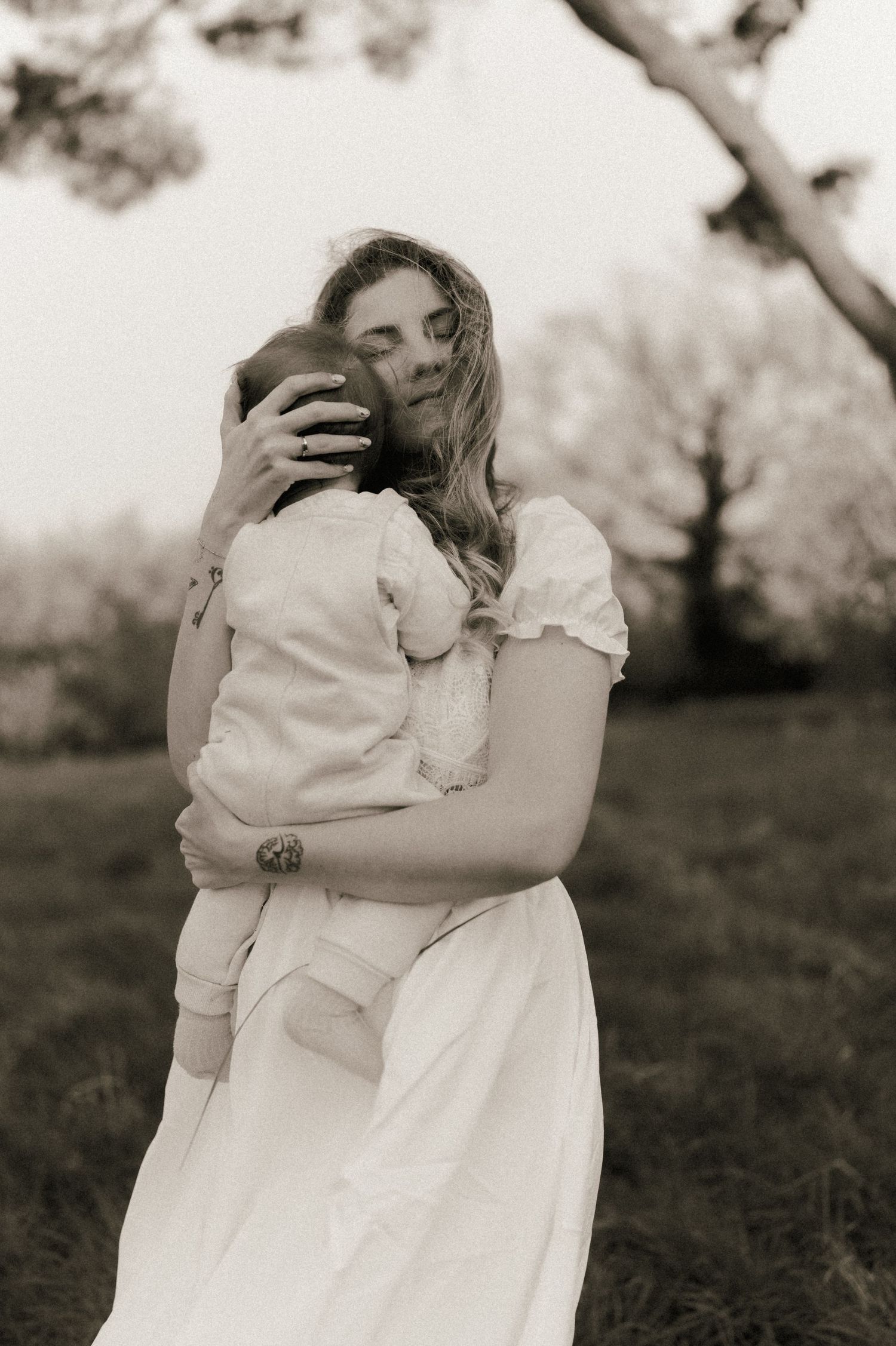 A mother in a white dress tenderly holds her baby outdoors in a serene black and white portrait.
