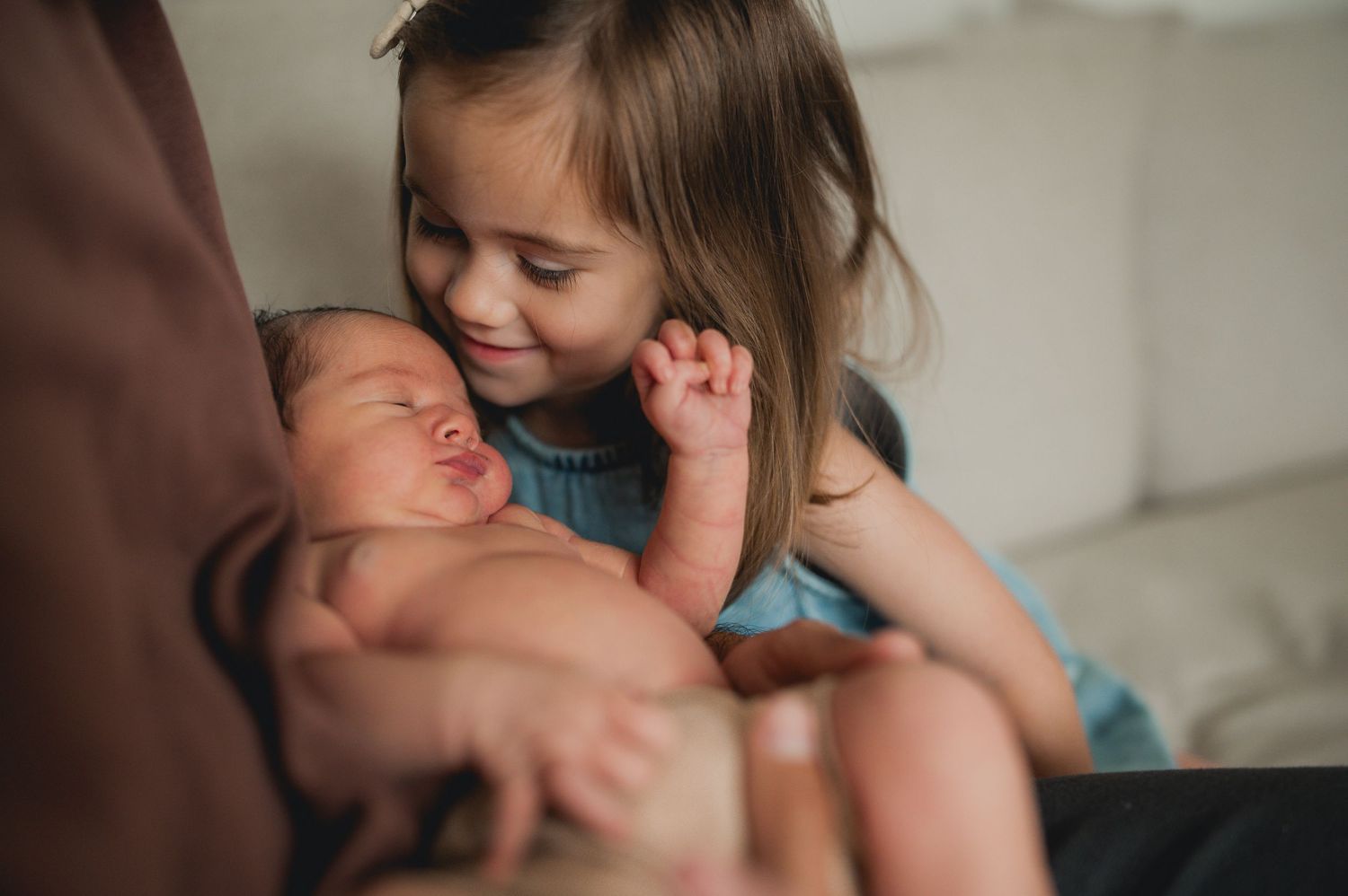 Older sister gently smiling and touching her newborn sibling during an indoor newborn photography session.