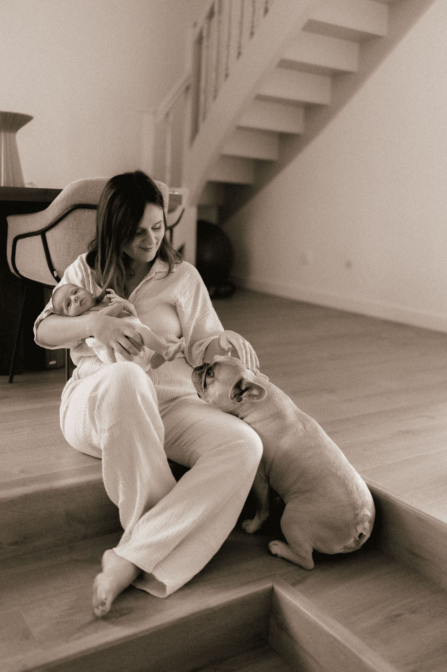 Mother in white outfit sitting on stairs holding newborn baby with a dog beside her, sepia tone.