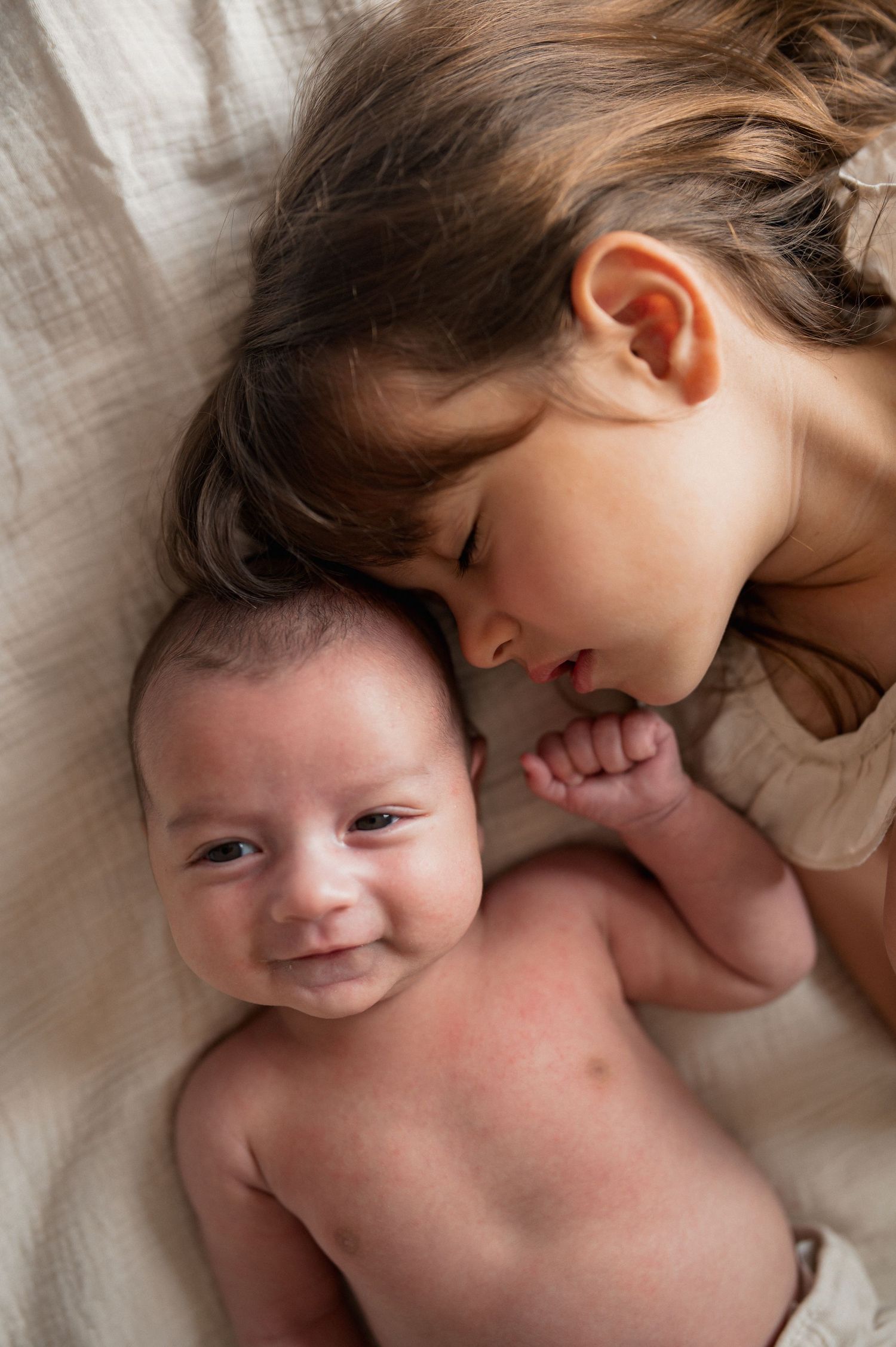 Adorable newborn baby smiling while older sibling leans in close, both lying on a soft beige blanket.