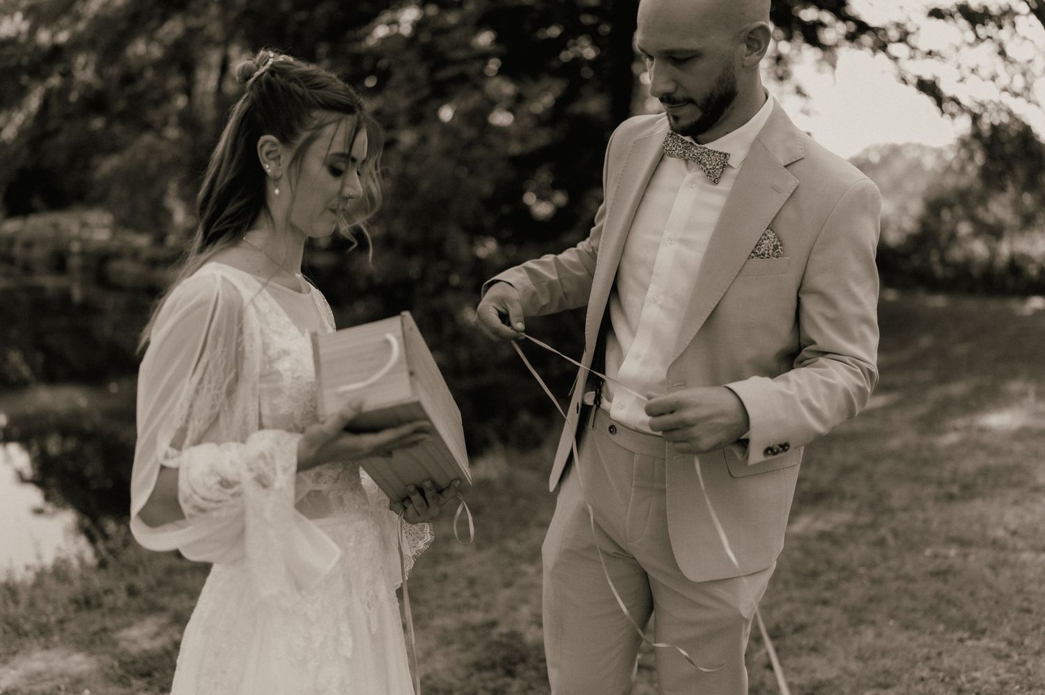 Black and white photo of a bride and groom exchanging vows outdoors, bride holding papers, groom in tan suit.