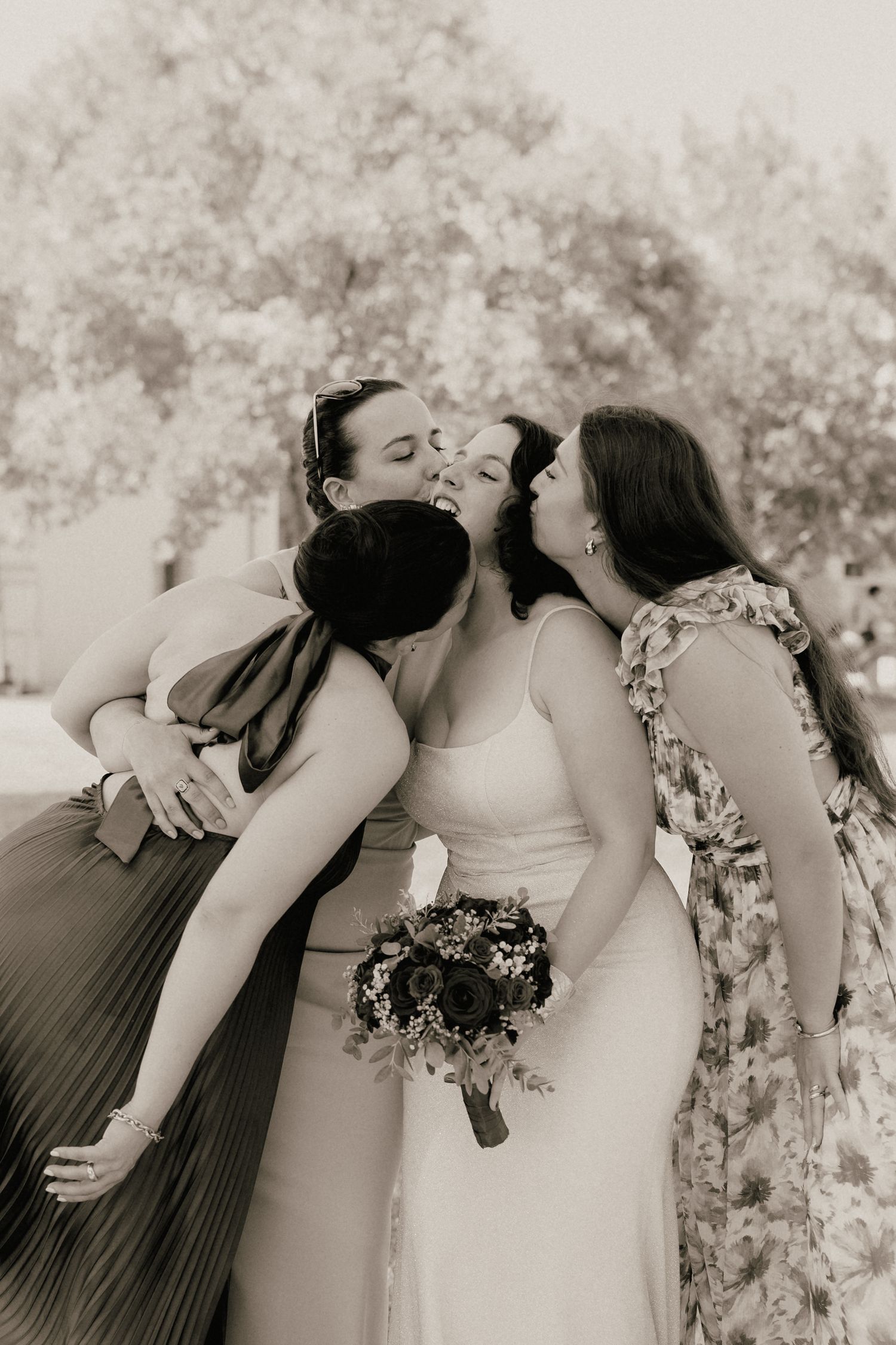Bride holding bouquet receives kisses on cheeks from two bridesmaids in black and white wedding photo outdoors.