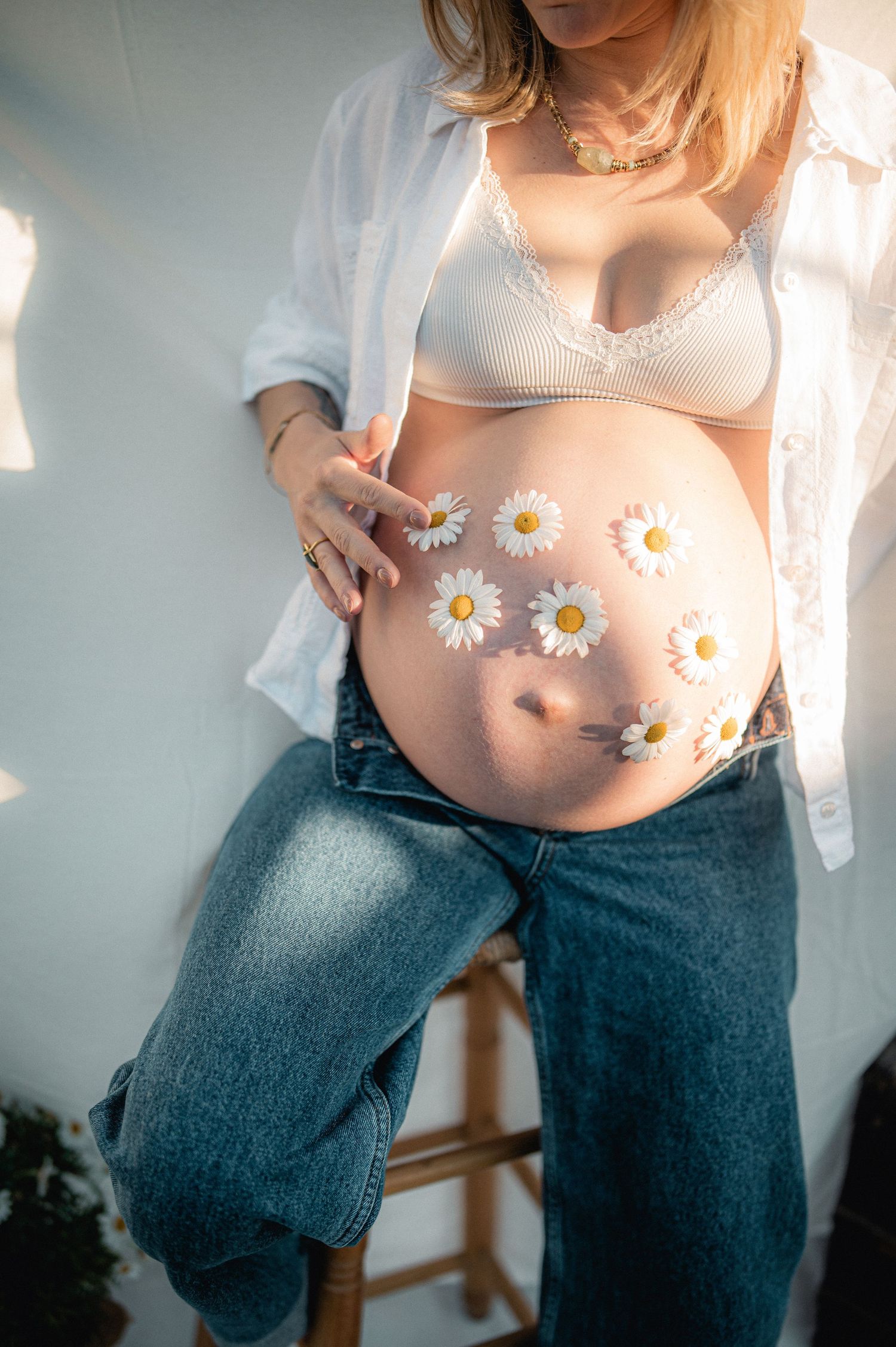 Pregnant belly decorated with white daisy flowers, wearing white bra and open denim jeans.