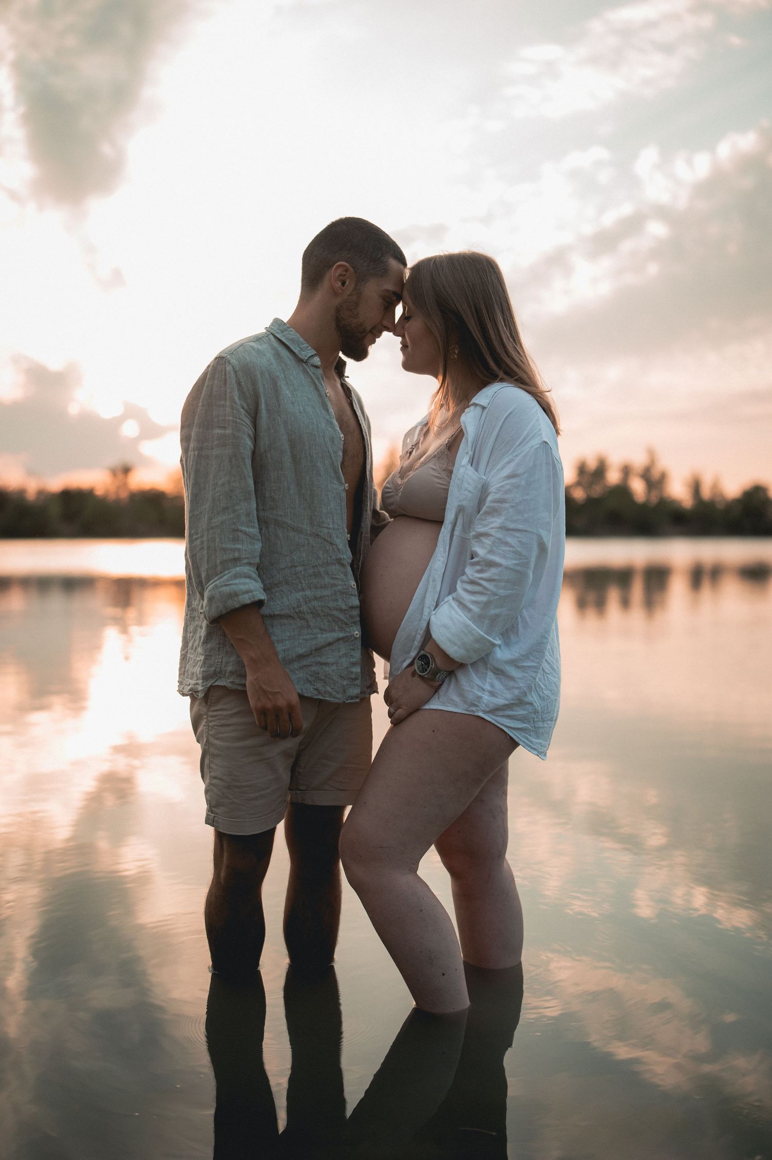 Expecting couple standing in shallow lake water touching foreheads at golden sunset.