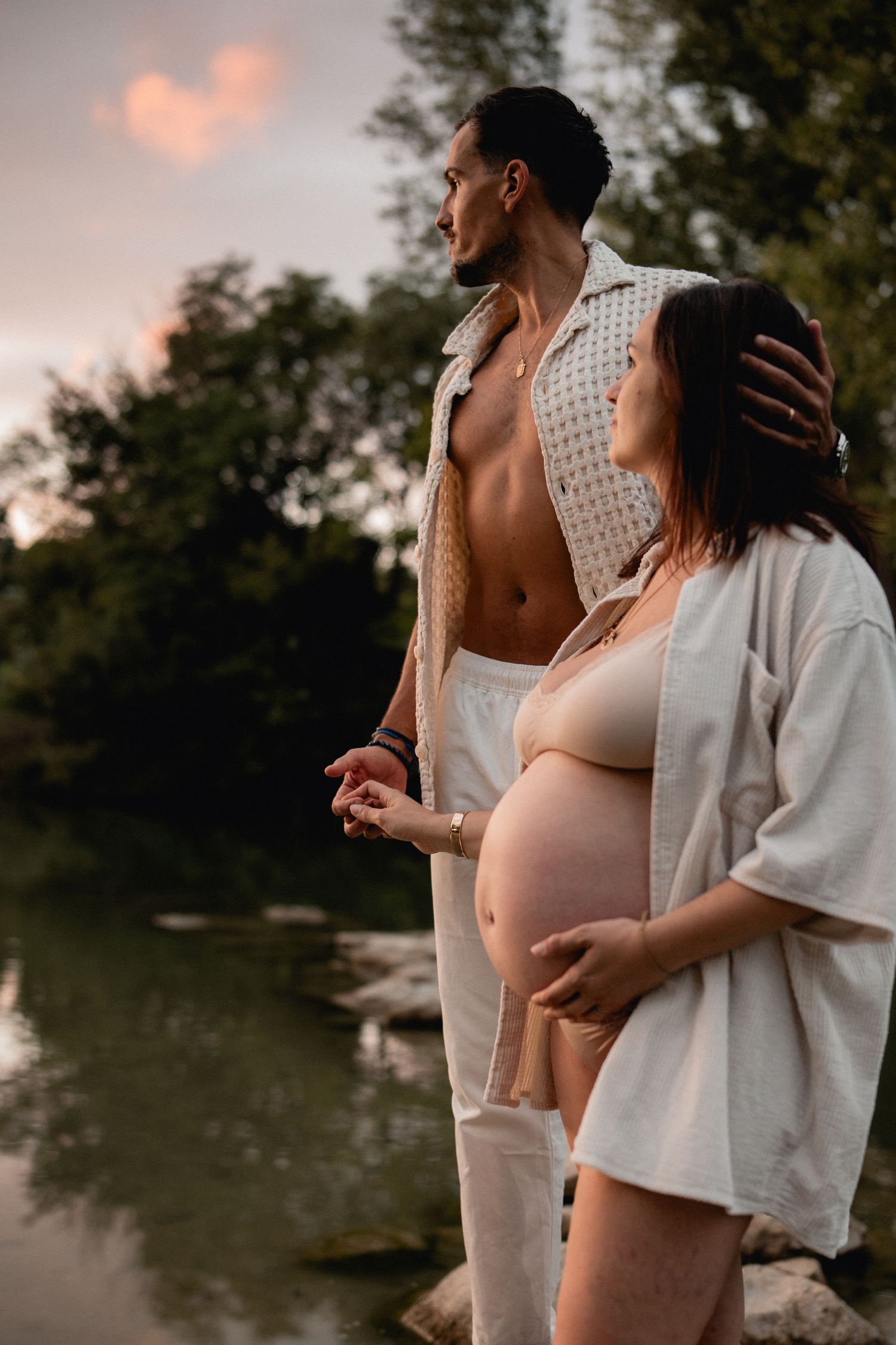 Pregnant couple standing by a serene river at sunset, woman cradling her baby bump in a romantic maternity photo.