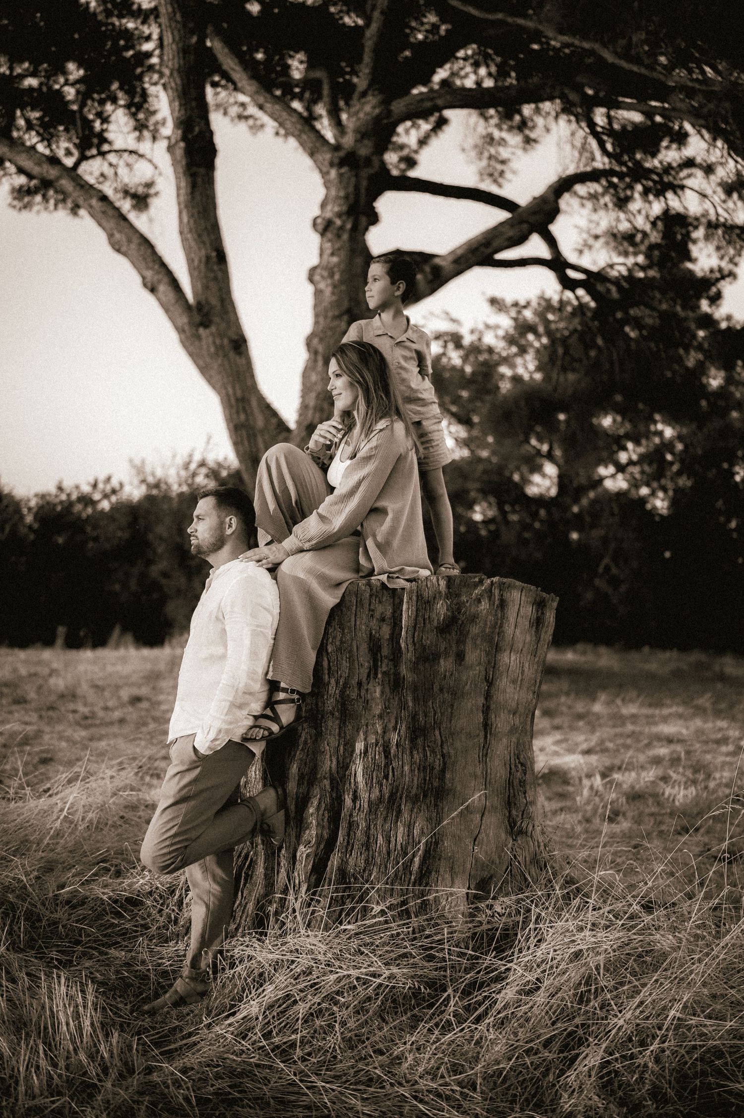Sepia-toned family portrait with child standing on tree stump above seated mother and resting father.