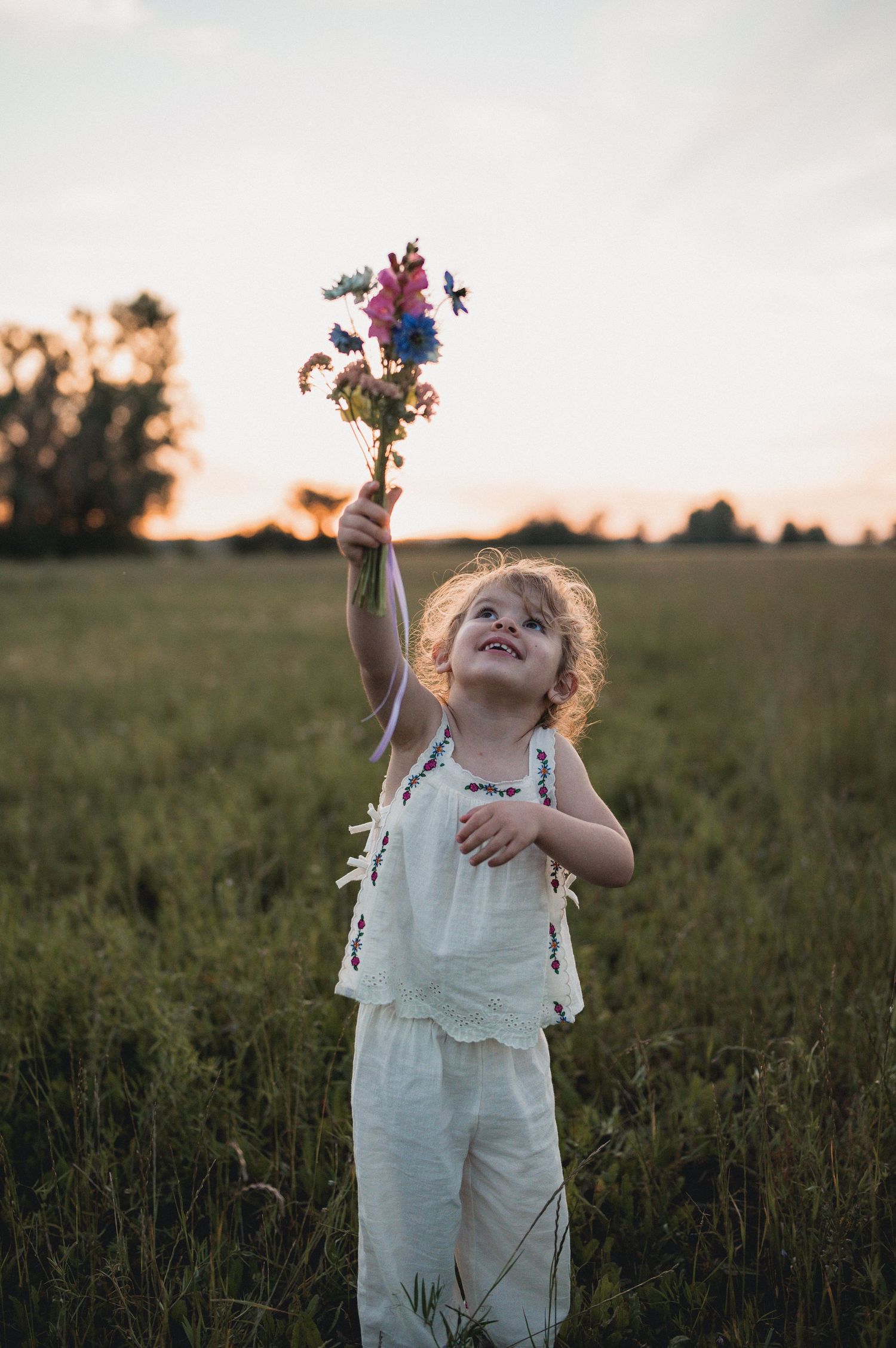 Happy toddler girl in white outfit holding wildflower bouquet in sunny green field at sunset.