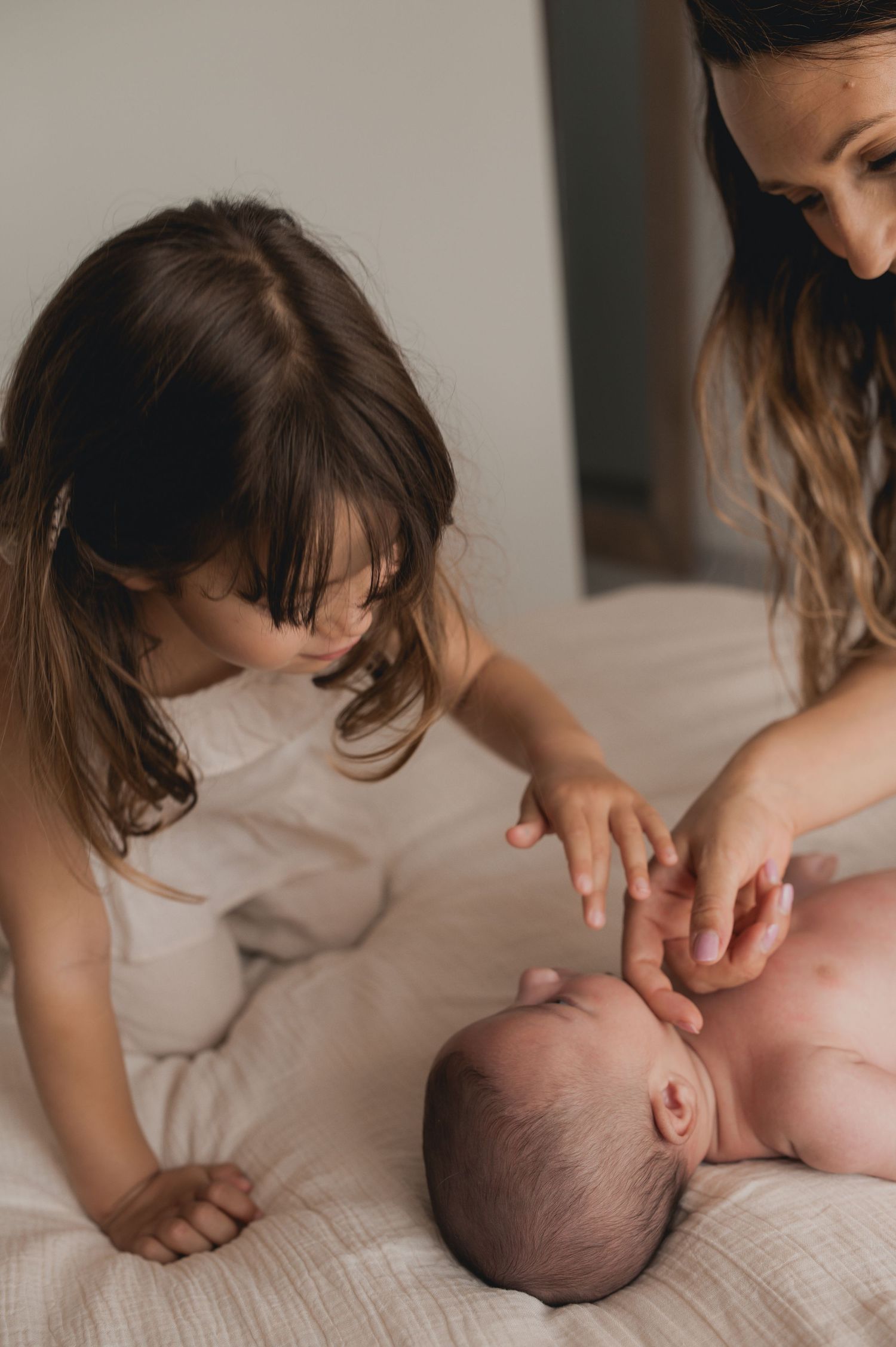 A mother and young daughter gently interact with a newborn baby lying on a white bed together.