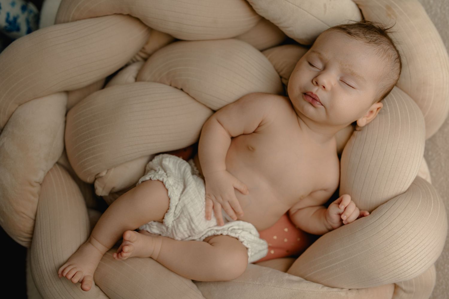 Newborn baby sleeping peacefully in a braided beige pillow nest wearing a white diaper.