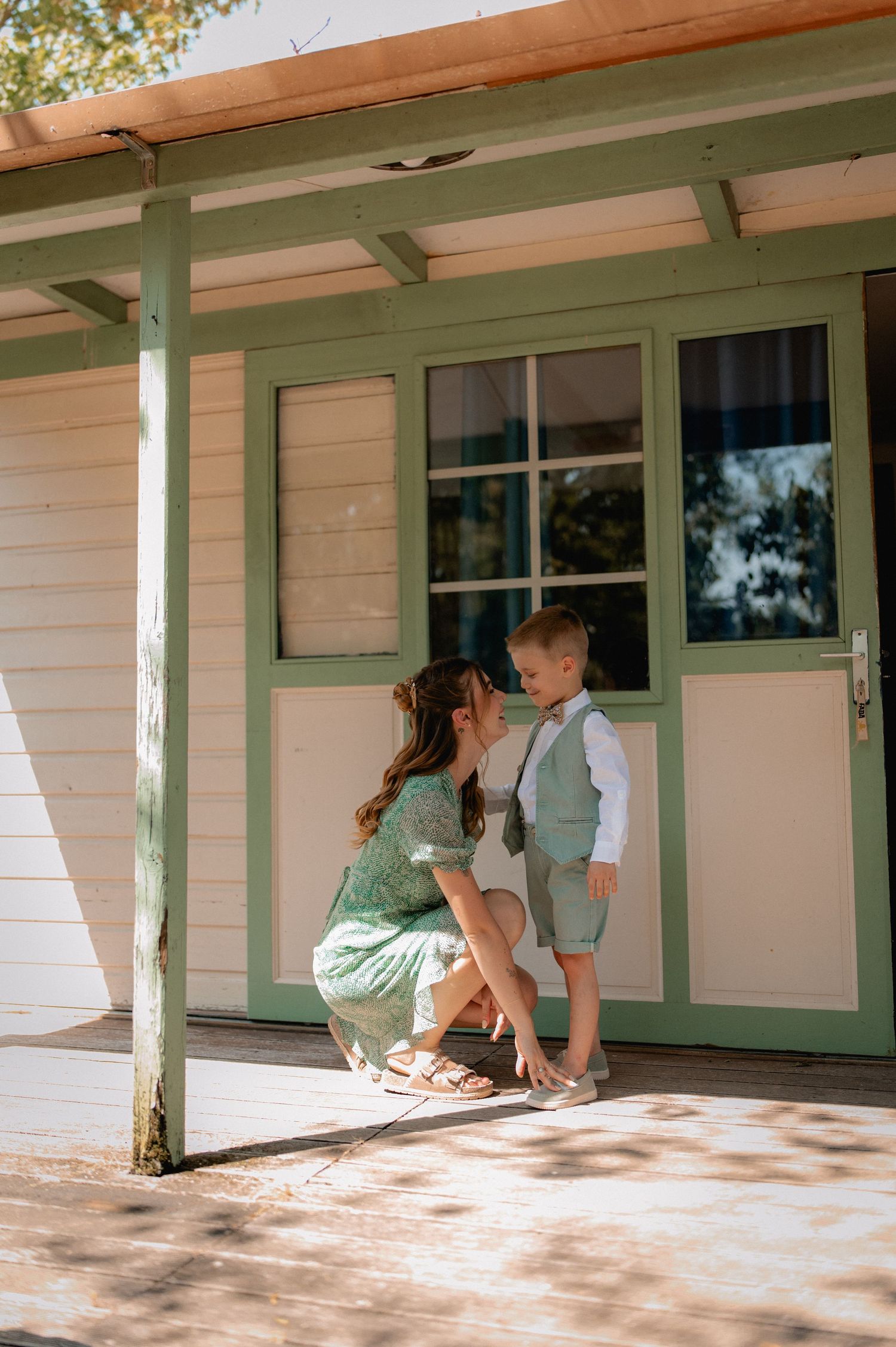 A mother crouches to kiss her young son in a green vest and shorts outside a charming white and green building.