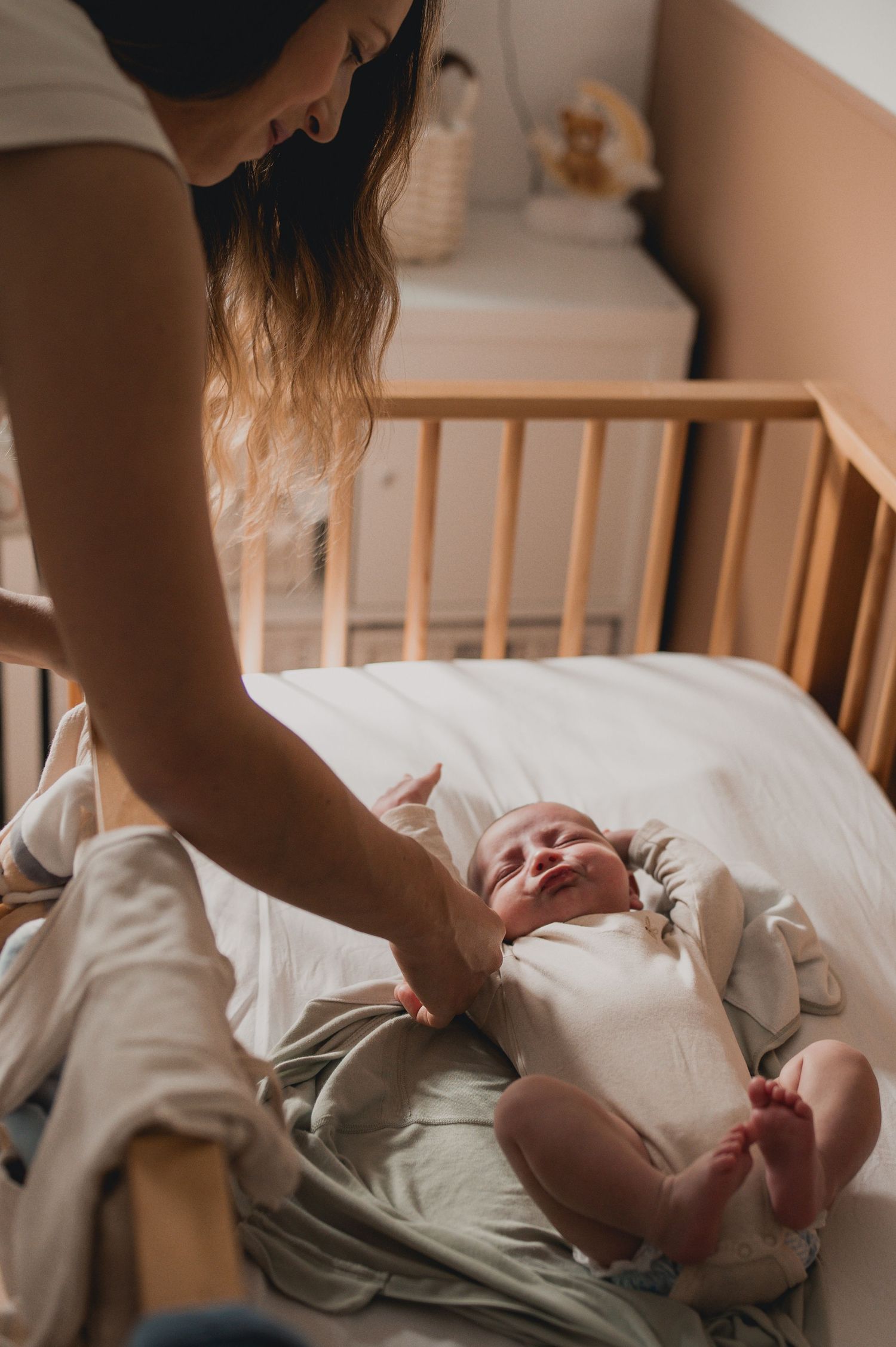 Mother smiling at her newborn baby lying in a wooden crib during an indoor lifestyle photo session.