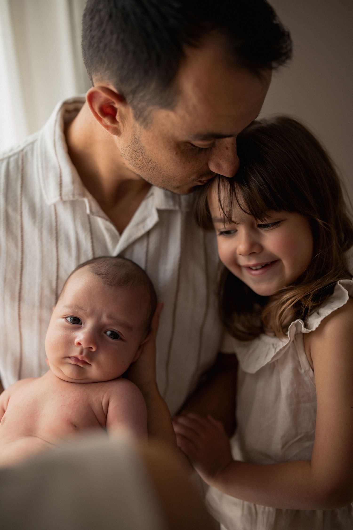 A loving father kisses his smiling toddler daughter while gently holding his newborn baby by a bright window.