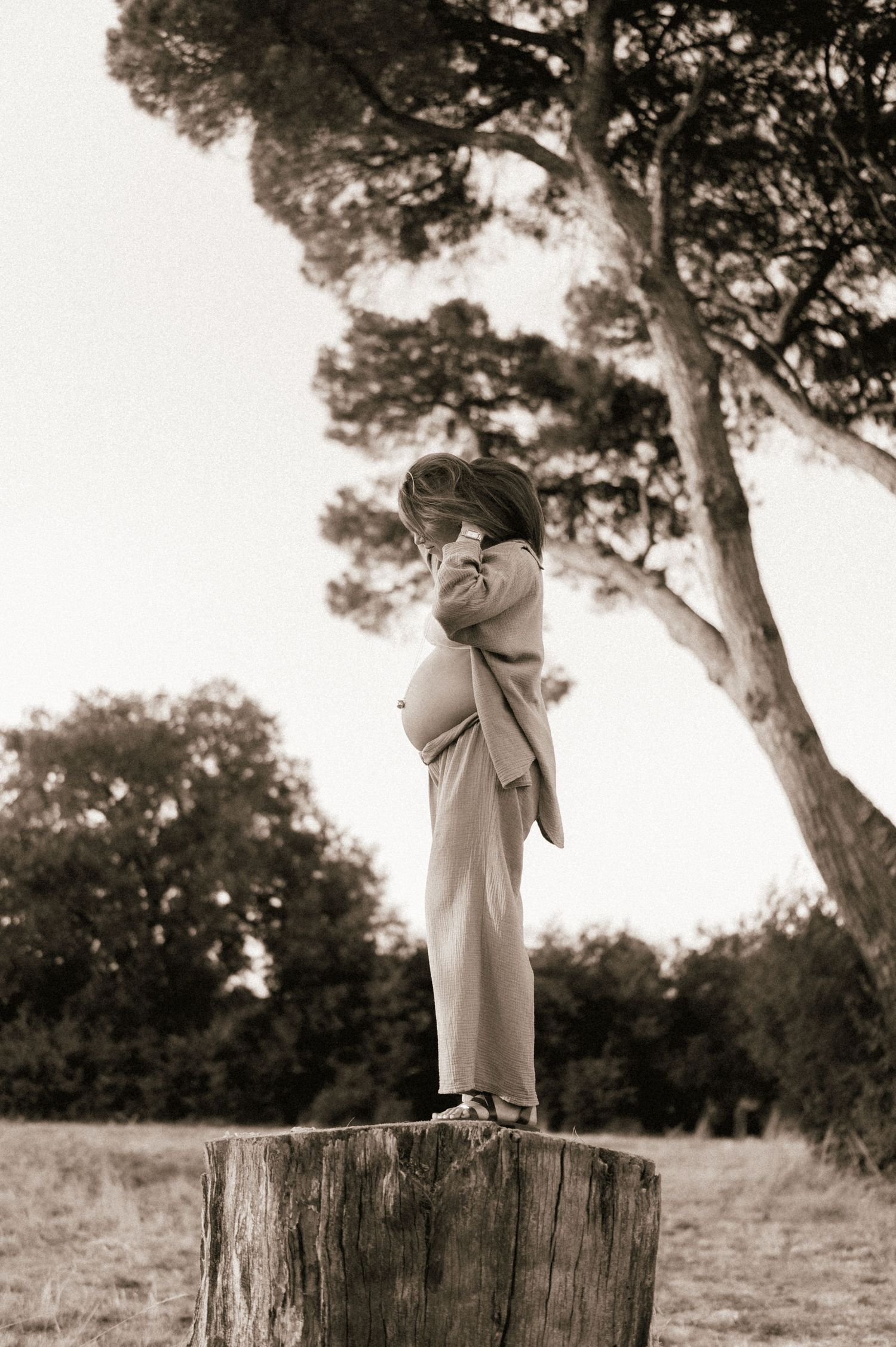 Sepia-toned photo of a pregnant woman standing on a tree stump in an outdoor field with tall trees.