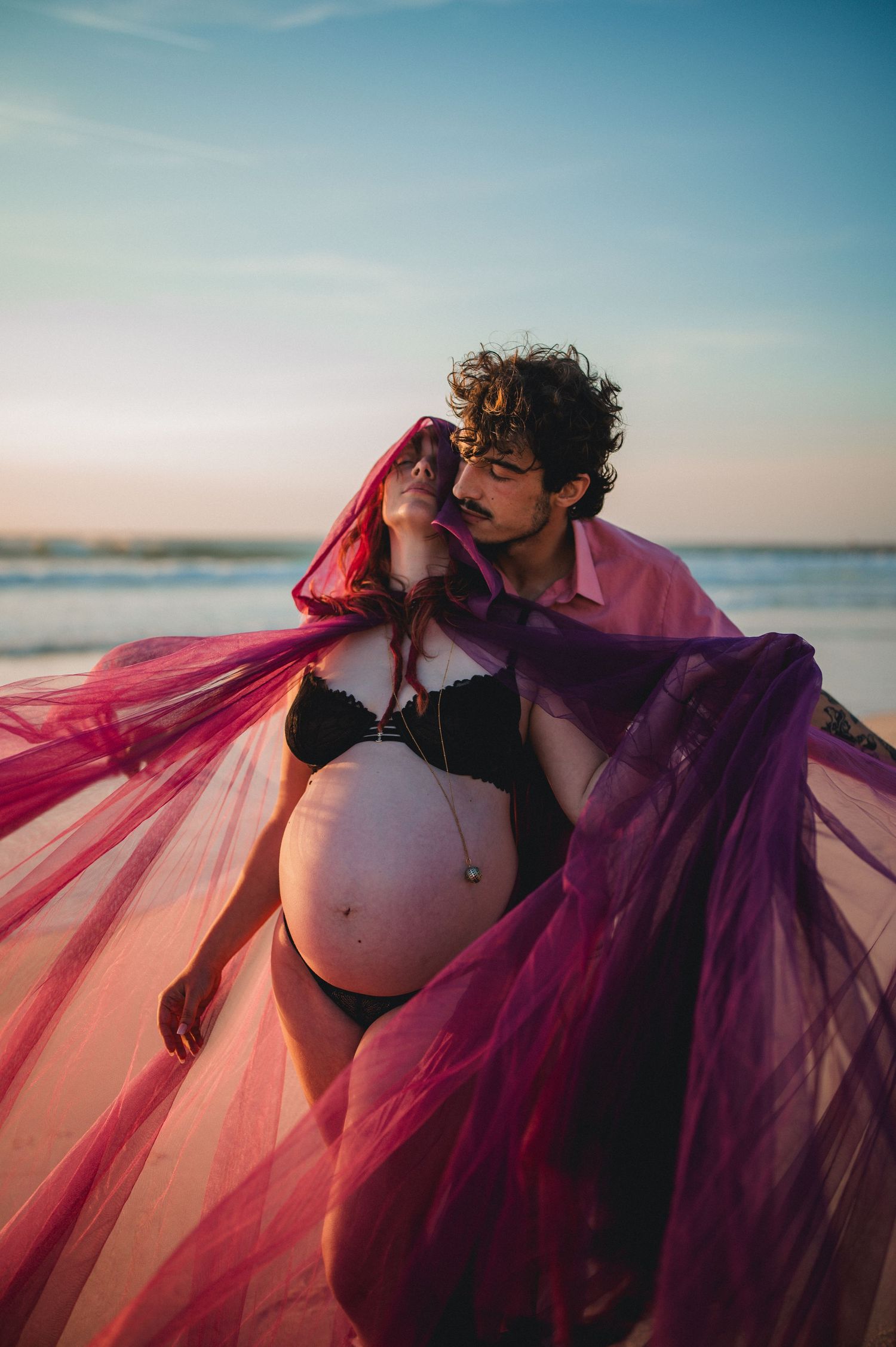 Pregnant woman in black bikini with flowing pink tulle fabric held by partner on beach at sunset.