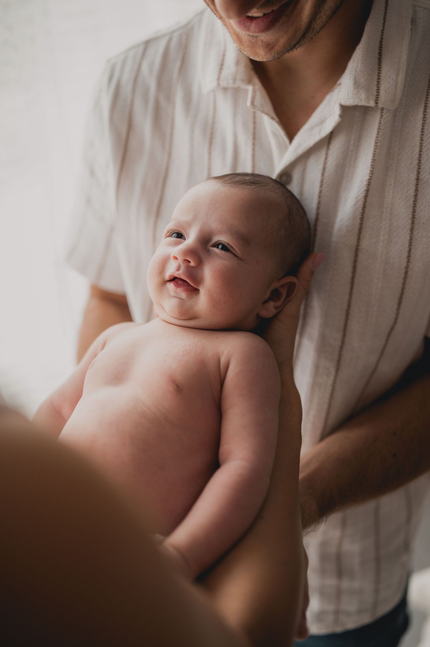 Smiling newborn baby being held by father in striped shirt near bright window.