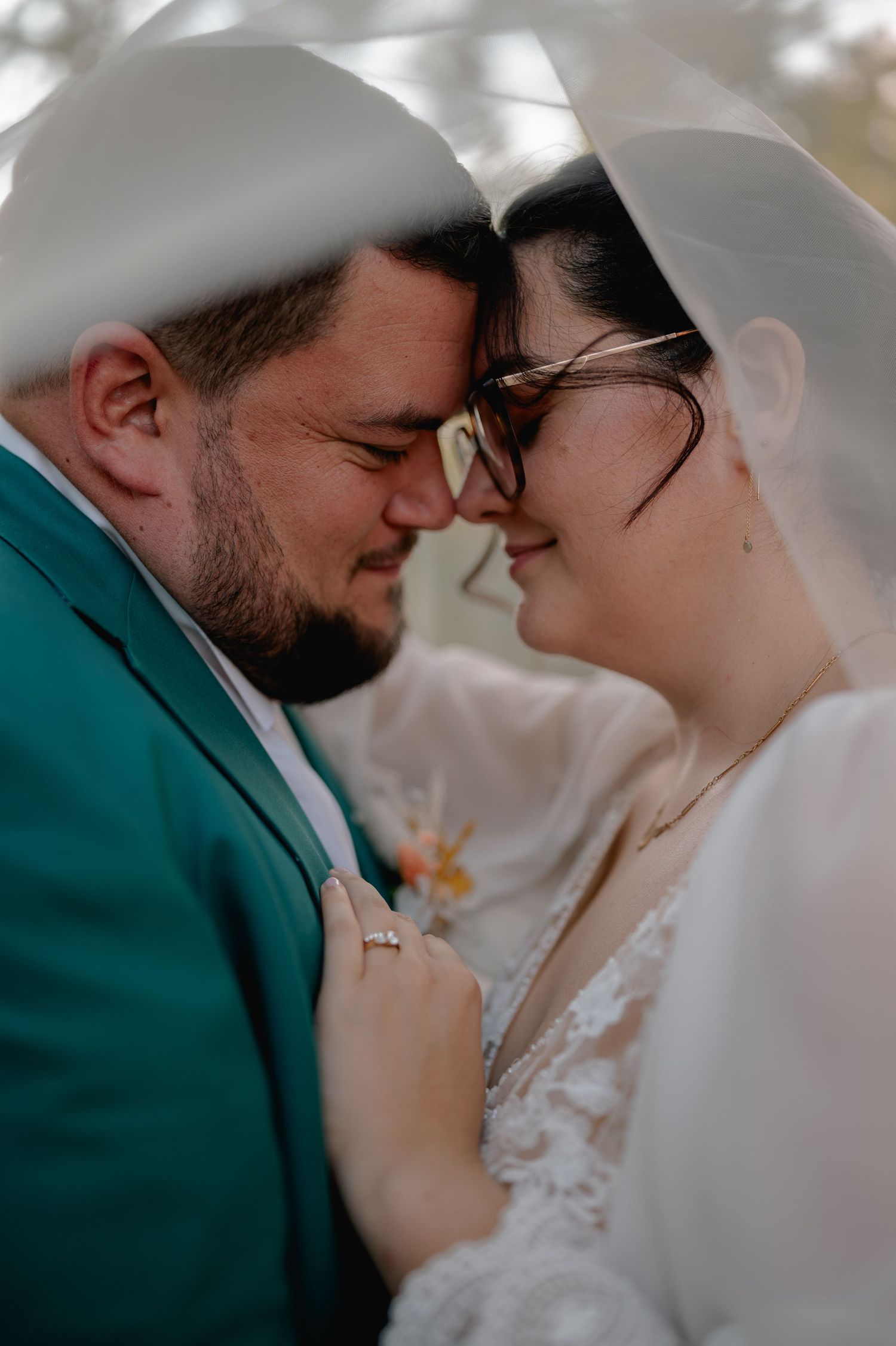 A bride and groom share an intimate forehead touch under a wedding veil, she wearing glasses and a lace dress, he in a teal suit.
