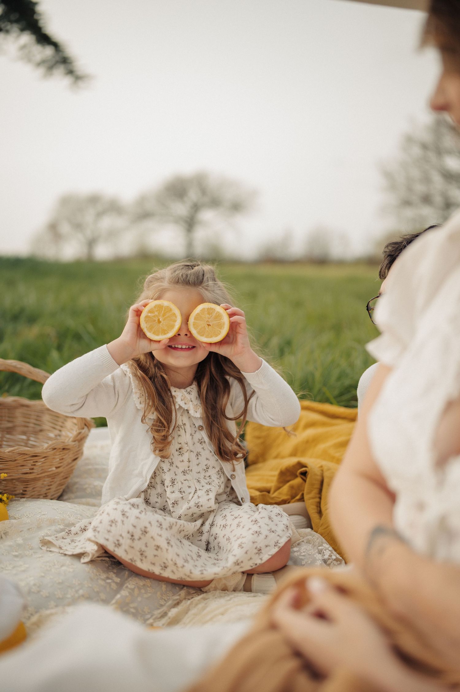 Smiling girl holding lemon slices over eyes at outdoor picnic on blanket in green field.
