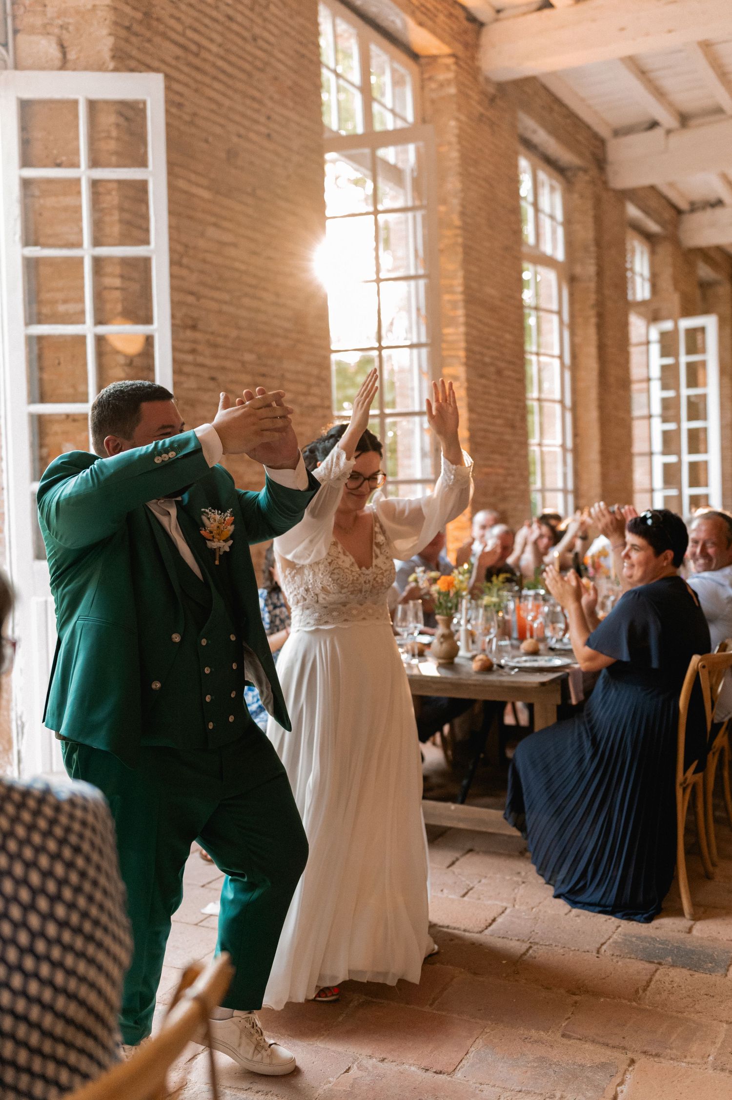 Bride and groom dancing joyfully at wedding reception in rustic brick venue, groom in green suit, guests cheering.