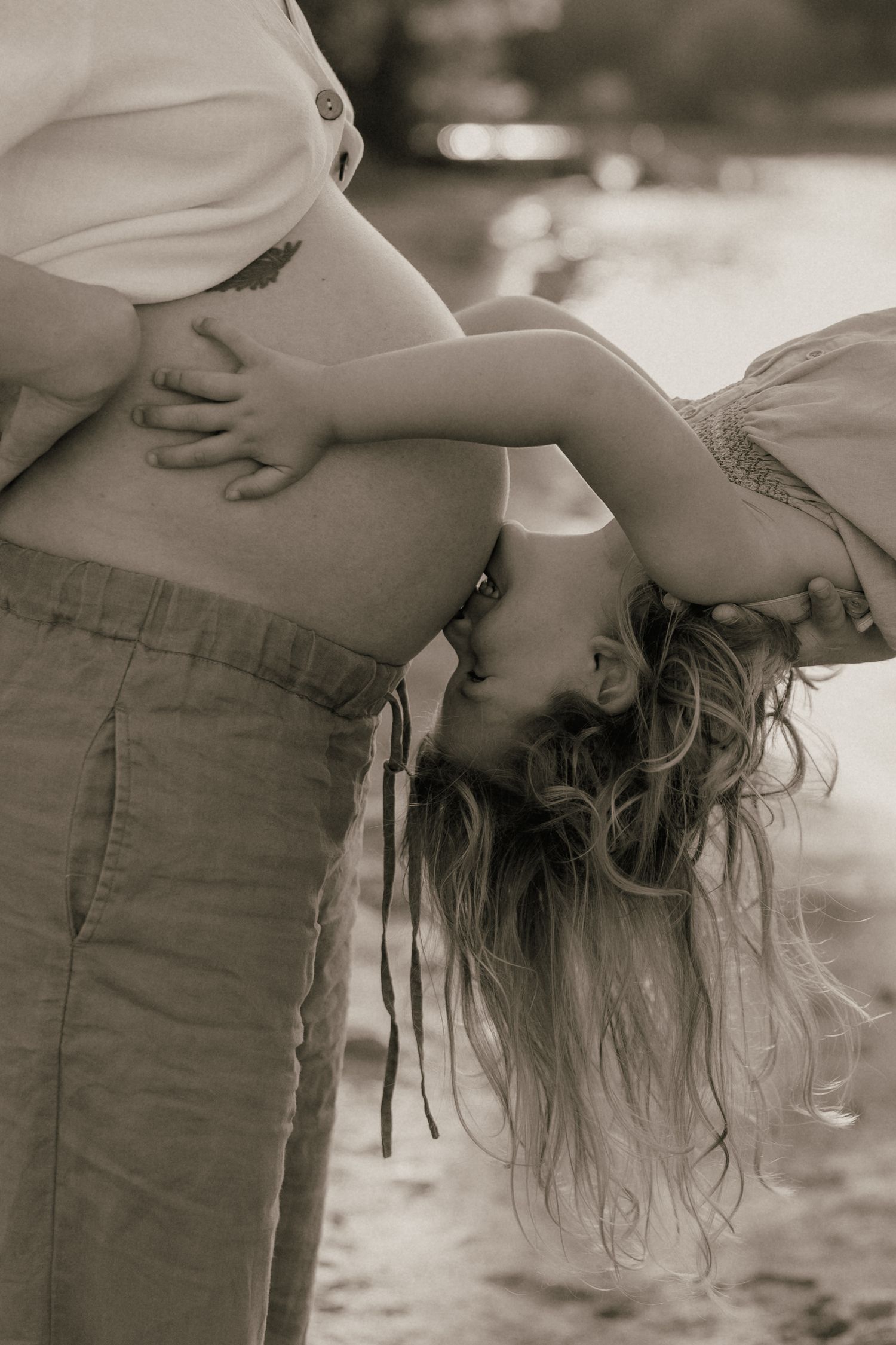 Black and white photo of child kissing pregnant mothers belly outdoors near water.