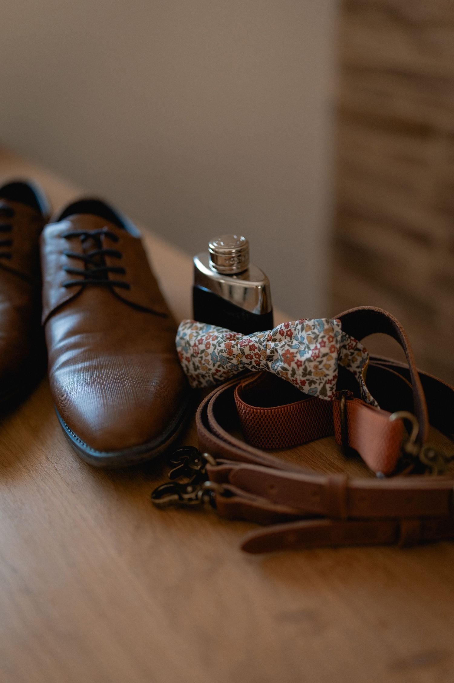 Brown leather dress shoes, floral bow tie, cologne bottle, belt, and suspenders arranged on a wooden surface.