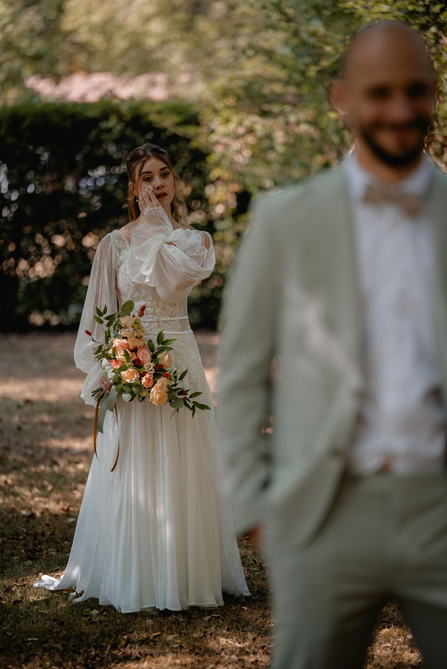 Bride in white gown with puff sleeves holds peach bouquet as groom in sage suit stands in foreground outdoors.