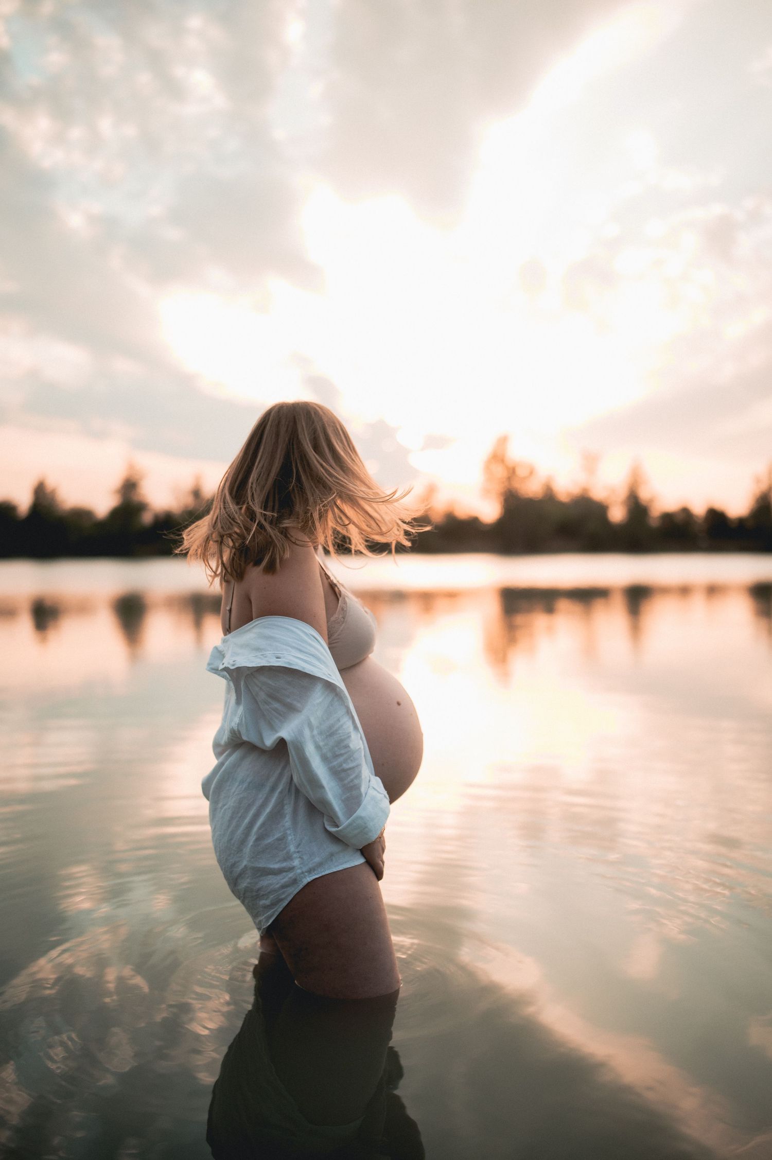 Pregnant woman standing in lake at golden sunset, white shirt open revealing baby bump.