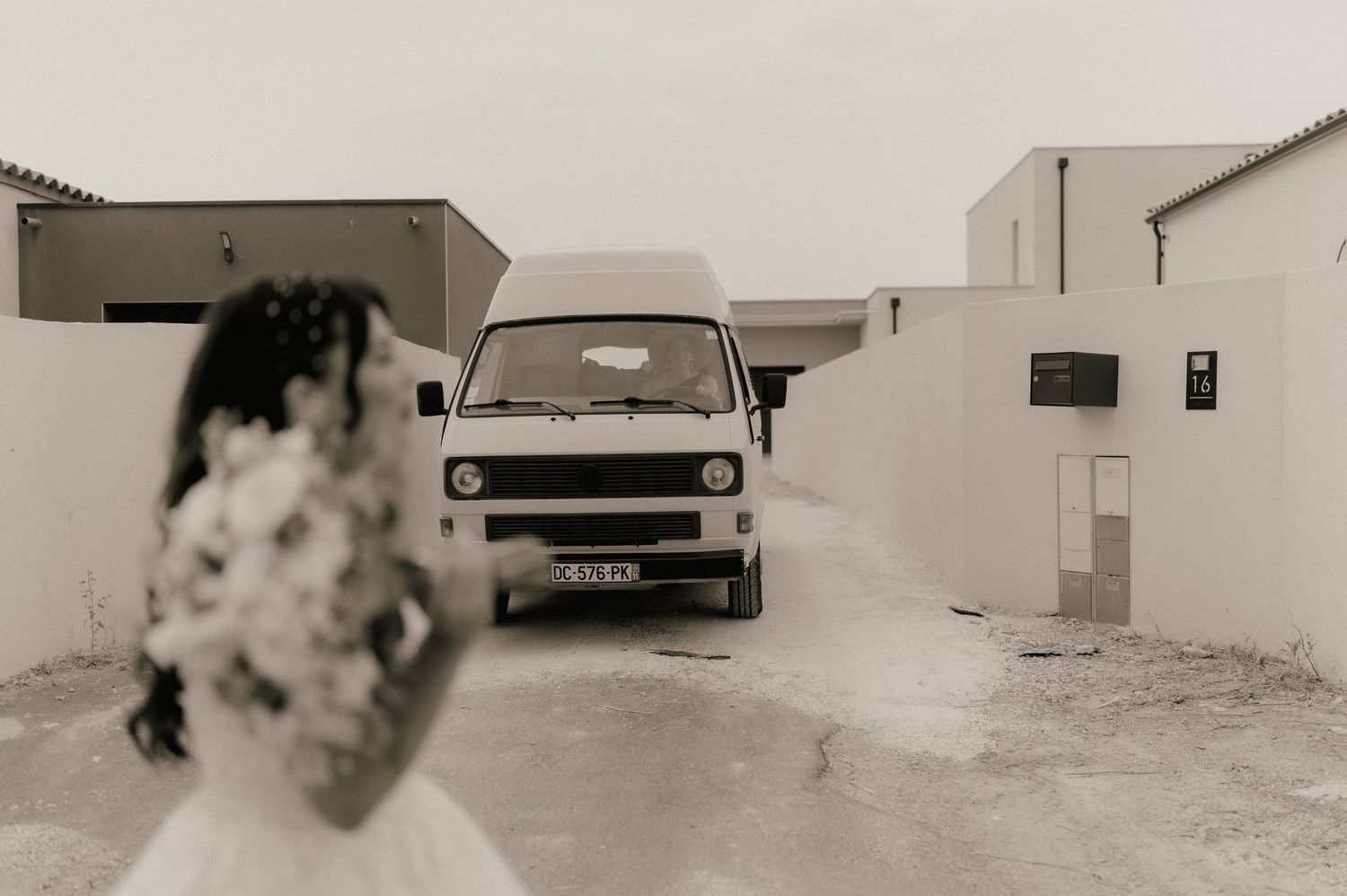 Bride in white gown holding bouquet stands before a vintage VW van in a black and white wedding photo.