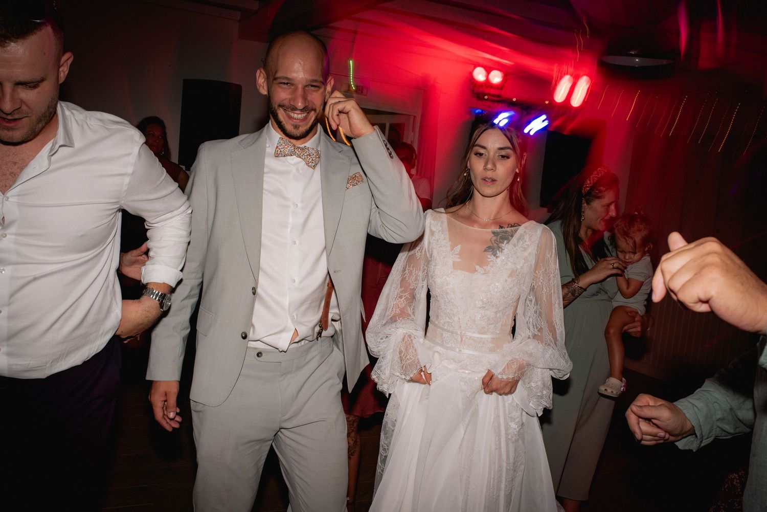 A joyful couple in wedding attire dancing at their reception, with red lighting and guests celebrating in the background.