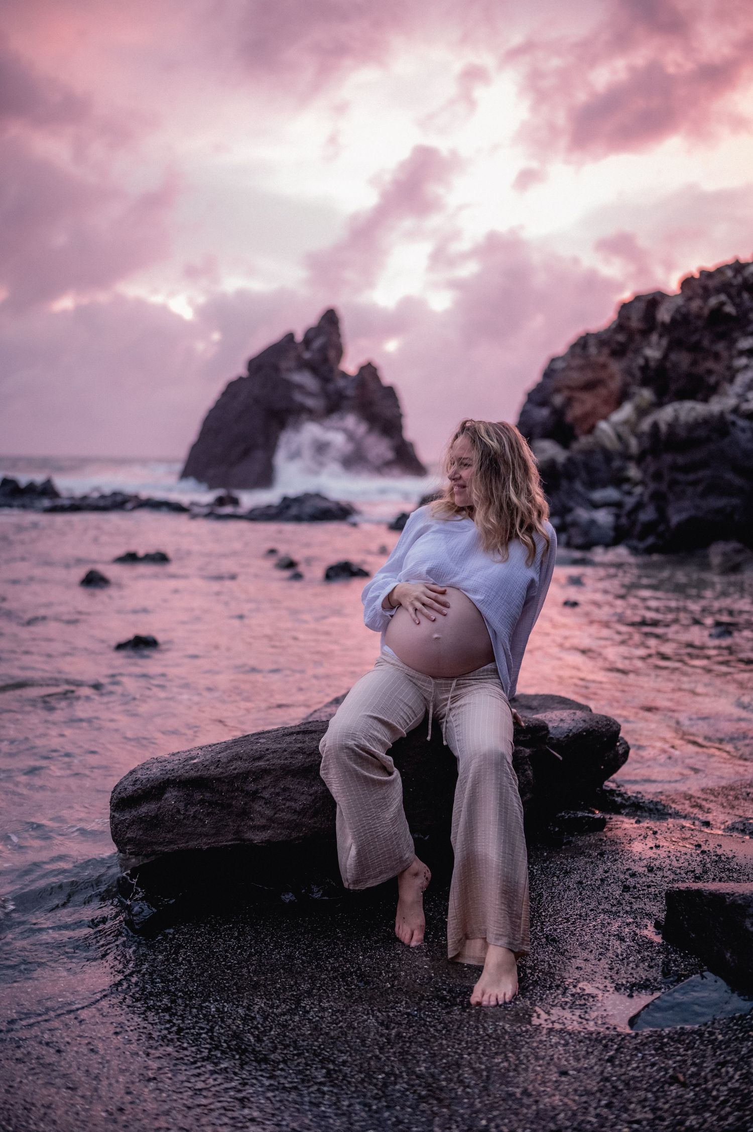 Pregnant woman sitting on coastal rocks cradling her belly at stunning pink sunset with ocean waves.
