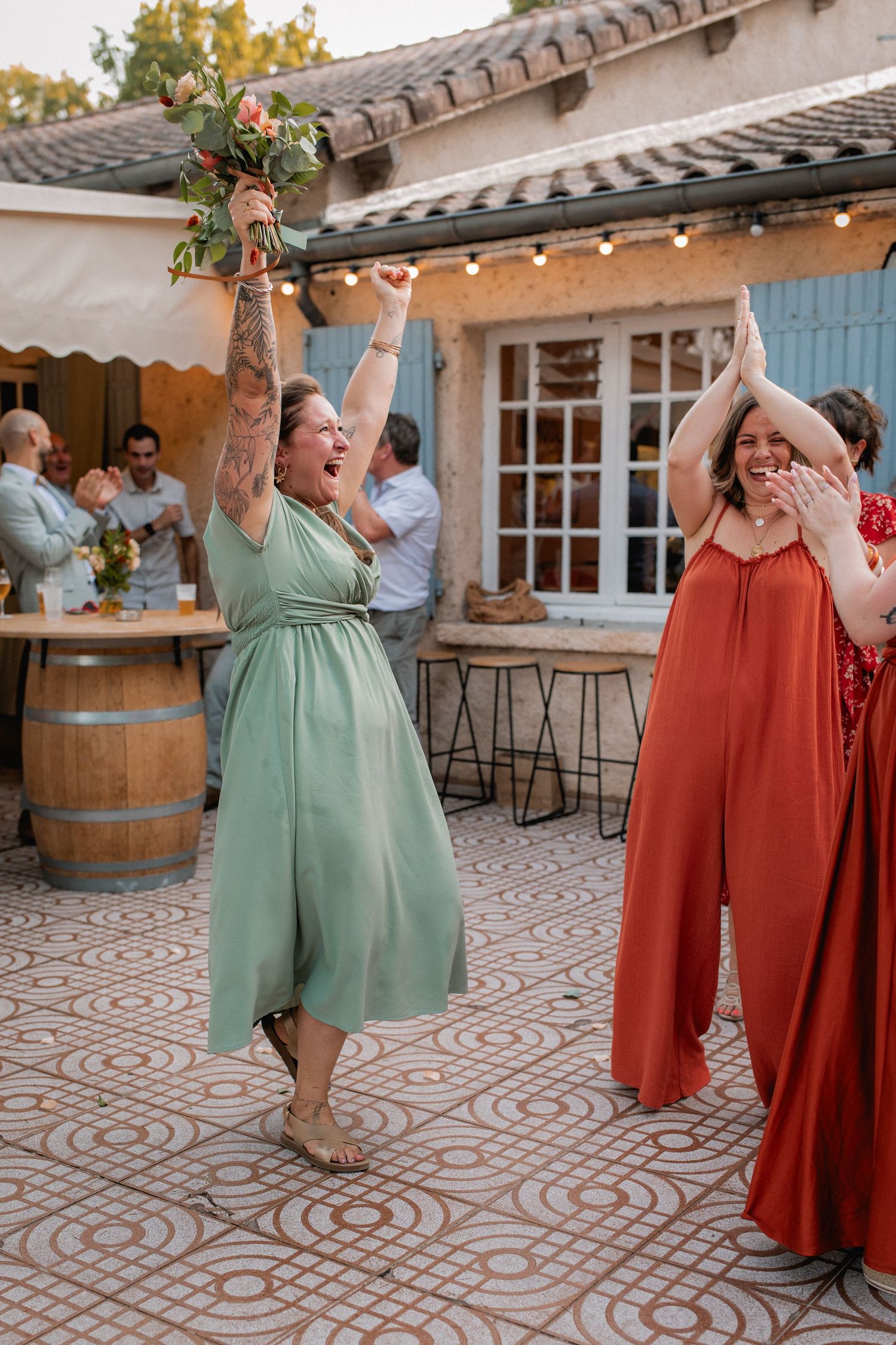 Woman in green dress catches wedding bouquet while another guest in red applauds at an outdoor reception.