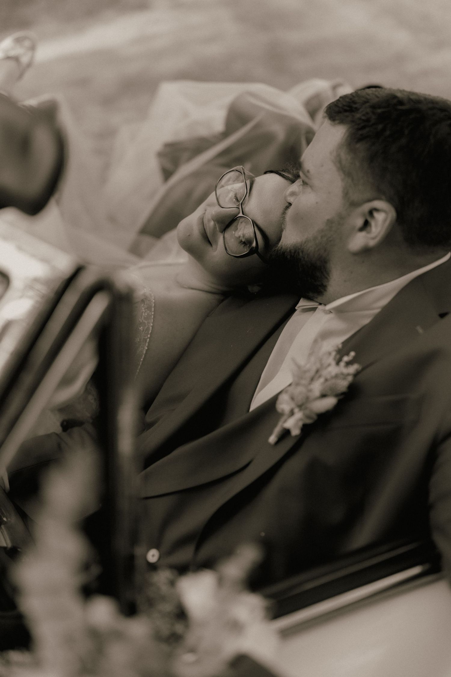 Bride and groom share tender moment in vintage car, black and white wedding photo.