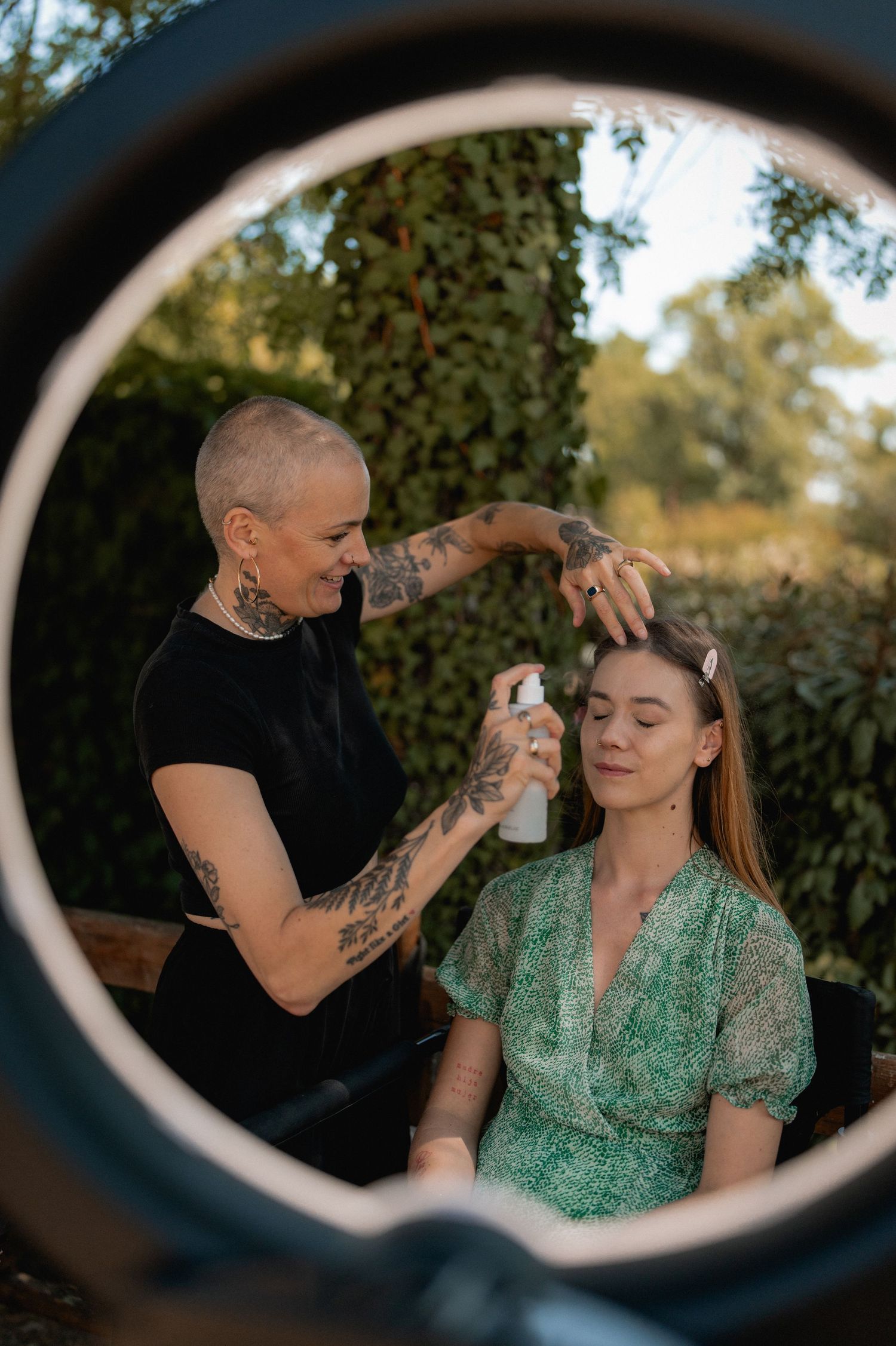 Tattooed hairstylist with shaved head sprays product on seated woman's hair outdoors near ivy-covered wall.